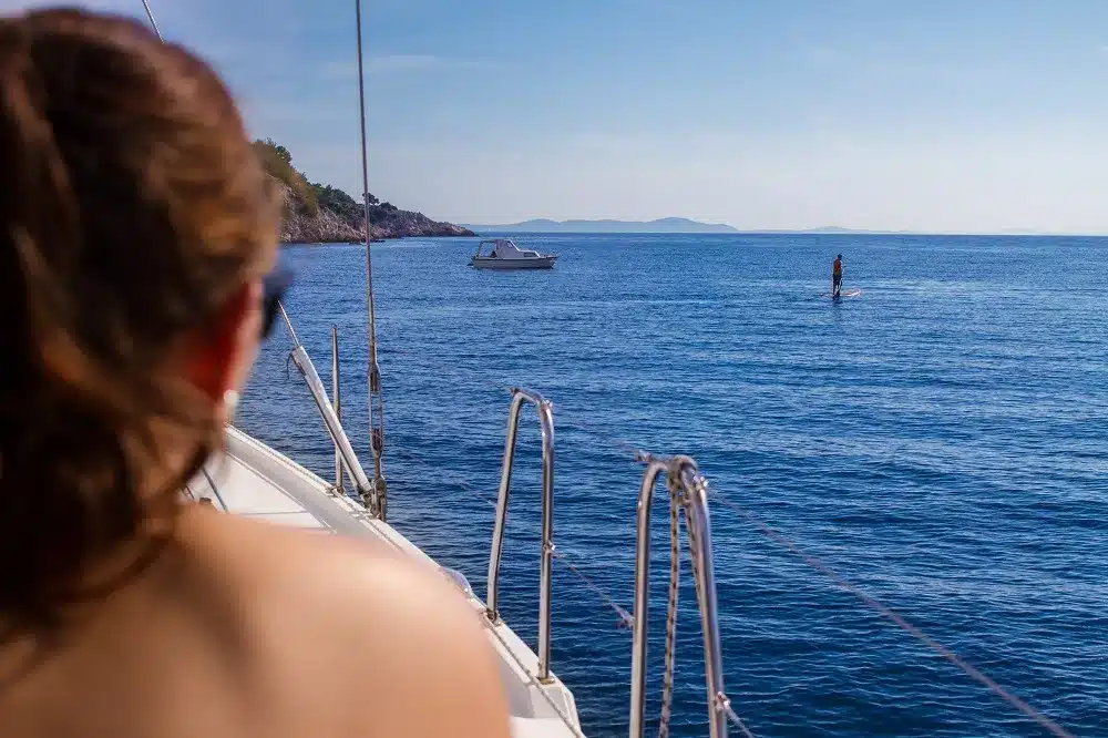 Eine Frau auf einem Boot blickt auf das offene Meer, wo in der Ferne unter einem klaren blauen Himmel ein weiteres Segelboot und eine Person auf einem Paddleboard zu sehen sind.