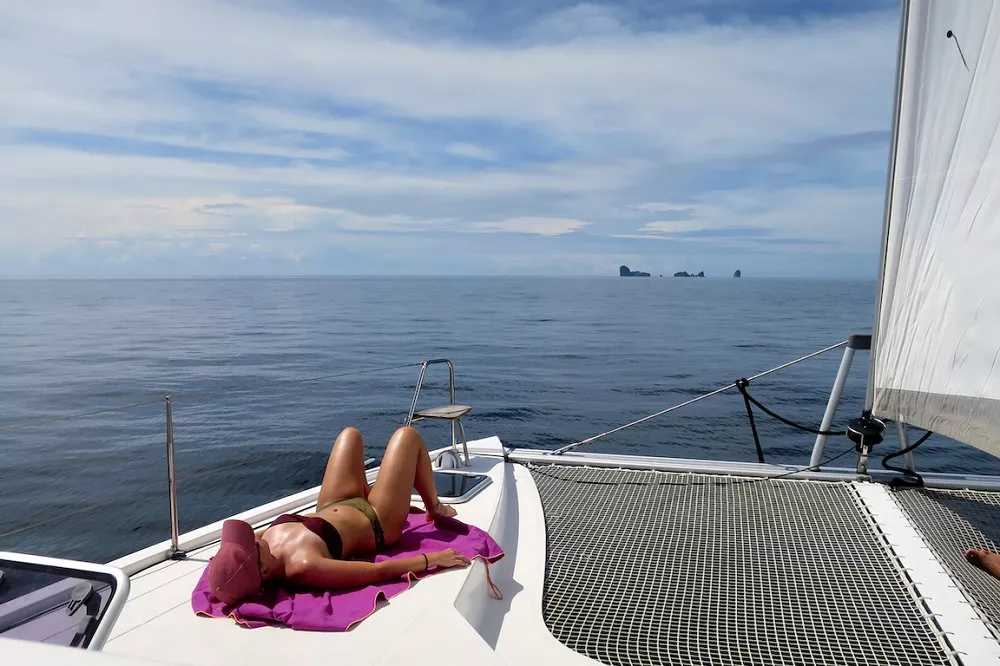 Eine Person sonnt sich während ihres Segelurlaubs auf dem Vorderdeck einer Segelyacht mit Blick auf das ruhige blaue Meer und die fernen Inseln unter einem klaren Himmel.