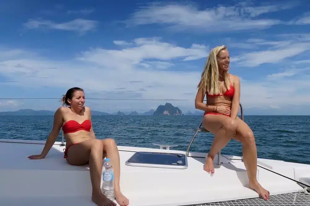 Zwei Frauen in roten Bikinis sitzen auf dem Bug eines Katamarans, lächeln einander an, vor der malerischen Kulisse des Ozeans und der Inseln unter einem klaren blauen Himmel.