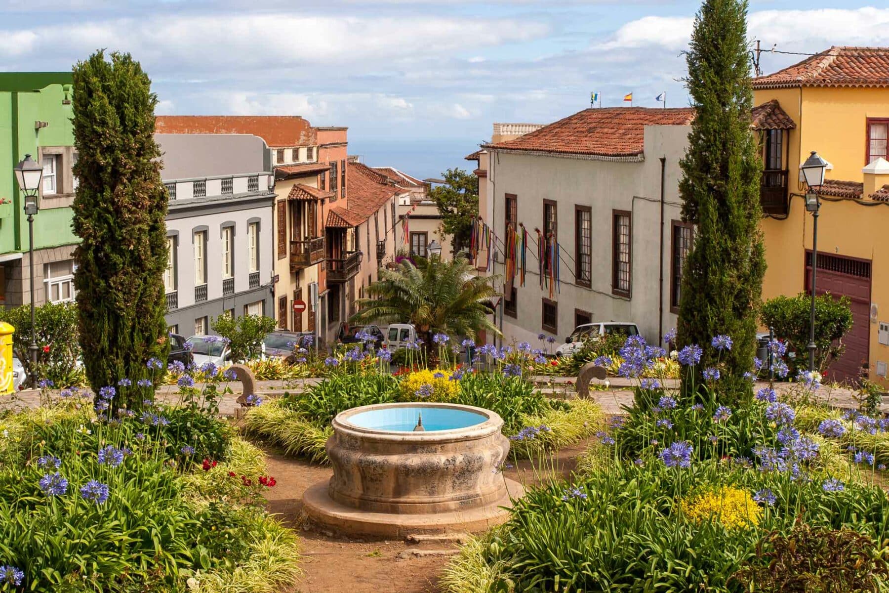 Eine malerische Straße in einer spanischen Stadt, gesäumt von traditionellen, farbenfrohen Gebäuden. Ein kleiner runder Brunnen steht in der Mitte eines Gartenbeets voller leuchtend violetter Blumen. Hohe Zypressen rahmen die Szene ein und geben den Blick frei auf das ferne Meer, auf dem Segelyachten anmutig unter einem teilweise bewölkten Himmel dahinsegeln.