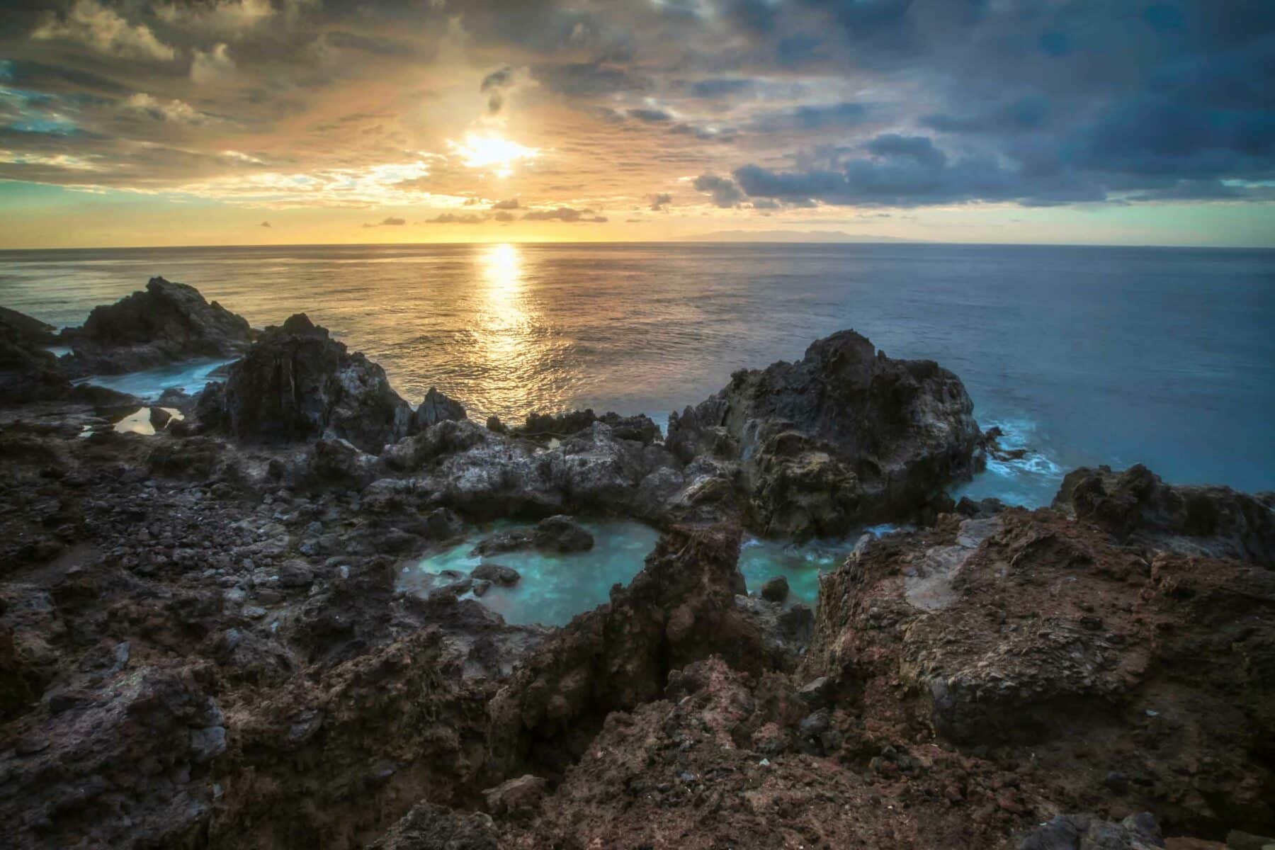 Sonnenuntergang über einer felsigen Küste mit Gezeitentümpeln während eines Segeltörns, bei dem die Sonne einen goldenen Schimmer auf die Wolken und das Meer wirft und die raue Textur der Felsen hervorhebt.