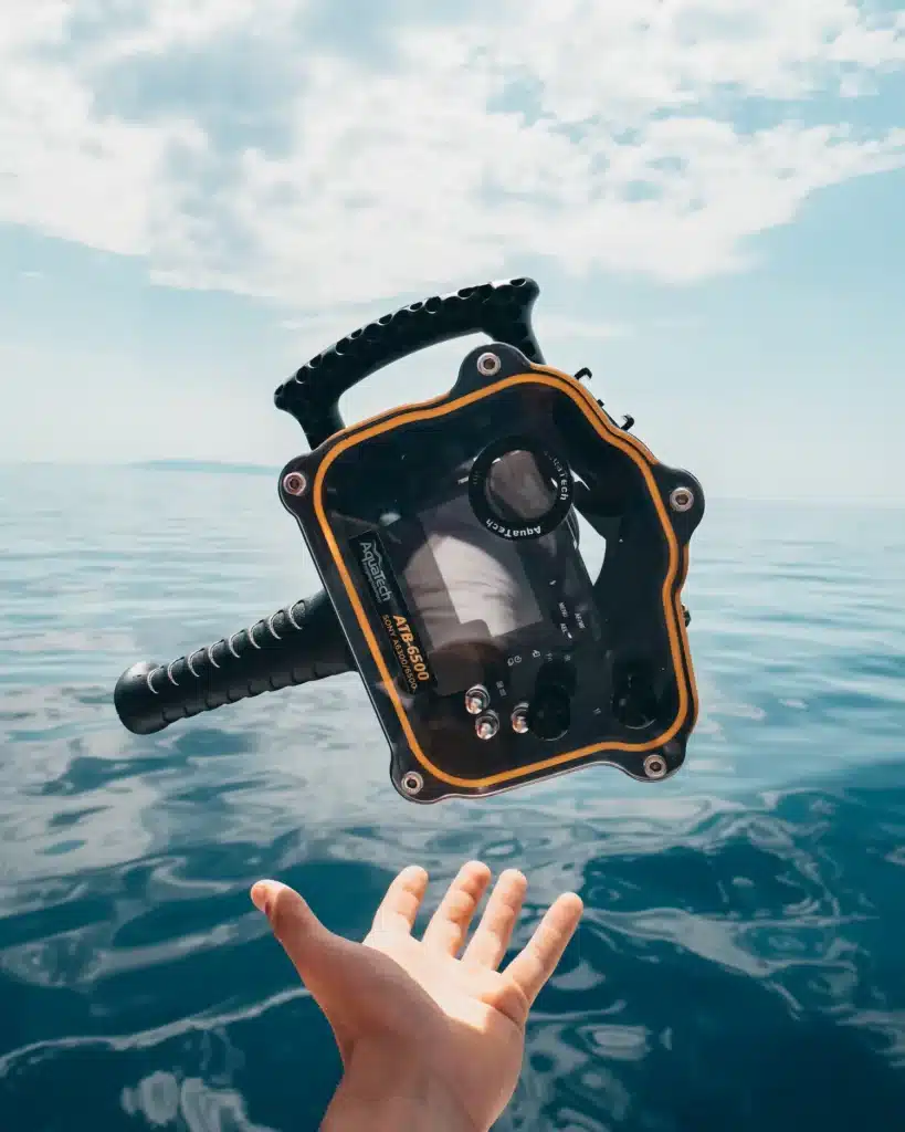 Eine ausgestreckte Hand versucht, während eines Segelurlaubs ein schwimmendes Unterwasserkameragehäuse mit einem klaren blauen Meer und Himmel im Hintergrund einzufangen.