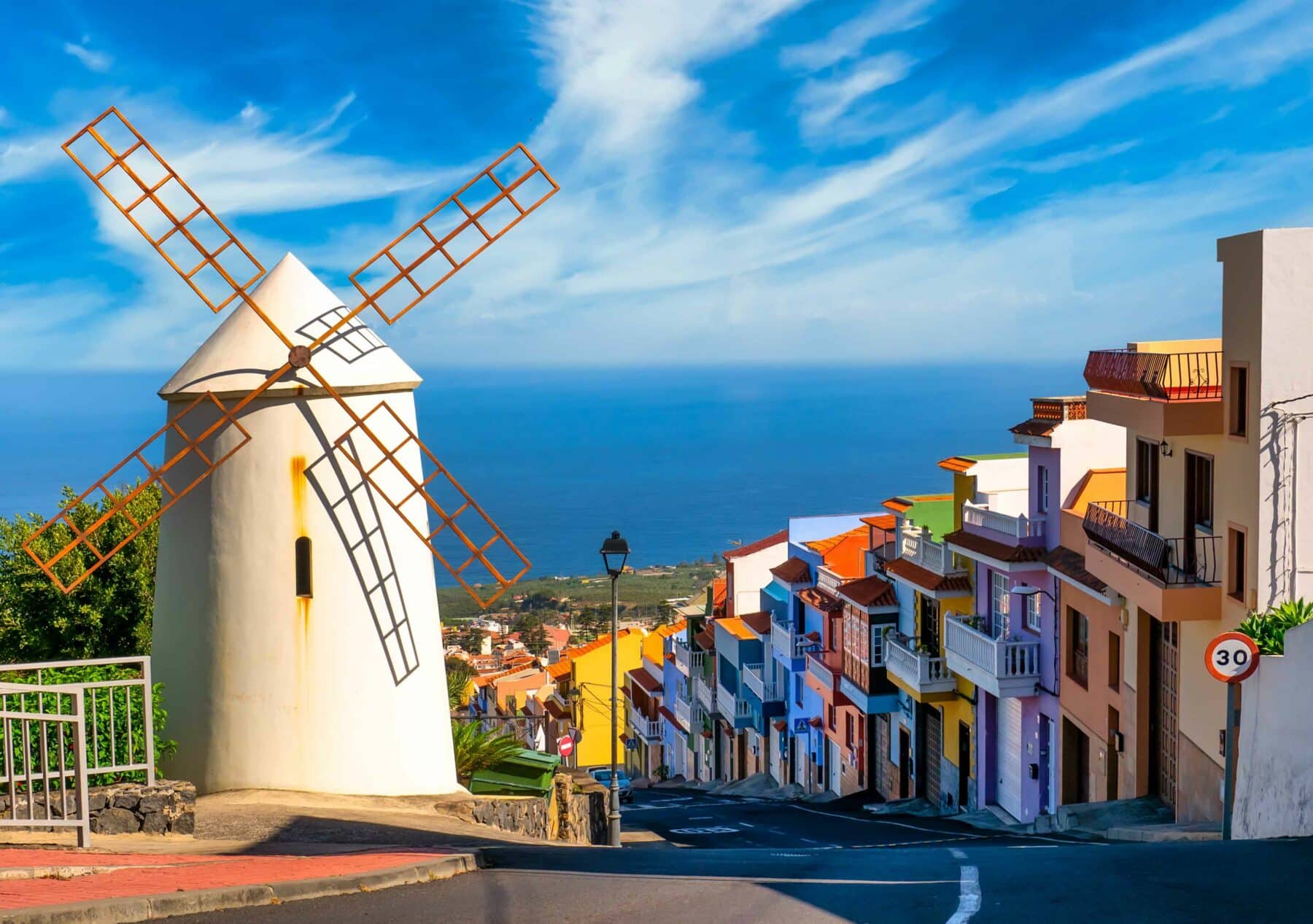 Eine malerische Aussicht auf eine Küstenstadt am Hang mit einer weißen Windmühle mit rostfarbenen Flügeln im Vordergrund. Bunt bemalte Häuser säumen eine gewundene Straße, die hinunter zum Meer führt, wo ein Katamaran auf einen abenteuerlichen Segeltörn wartet. Der Himmel ist blau mit dünnen Wolken und ein Geschwindigkeitsbegrenzungsschild von 30 km/h ist sichtbar.