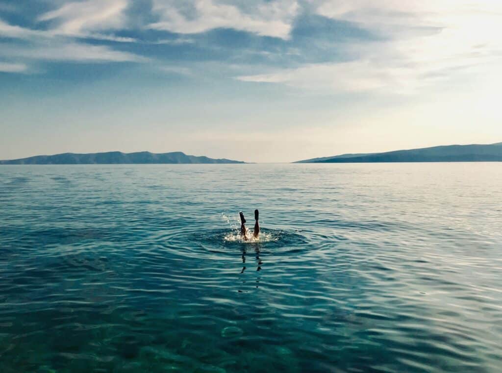 Eine Person taucht in das klare, blaue Wasser eines ruhigen Meeres ein, mit entfernten Hügeln und einem Katamaran unter einem klaren Himmel im Hintergrund.