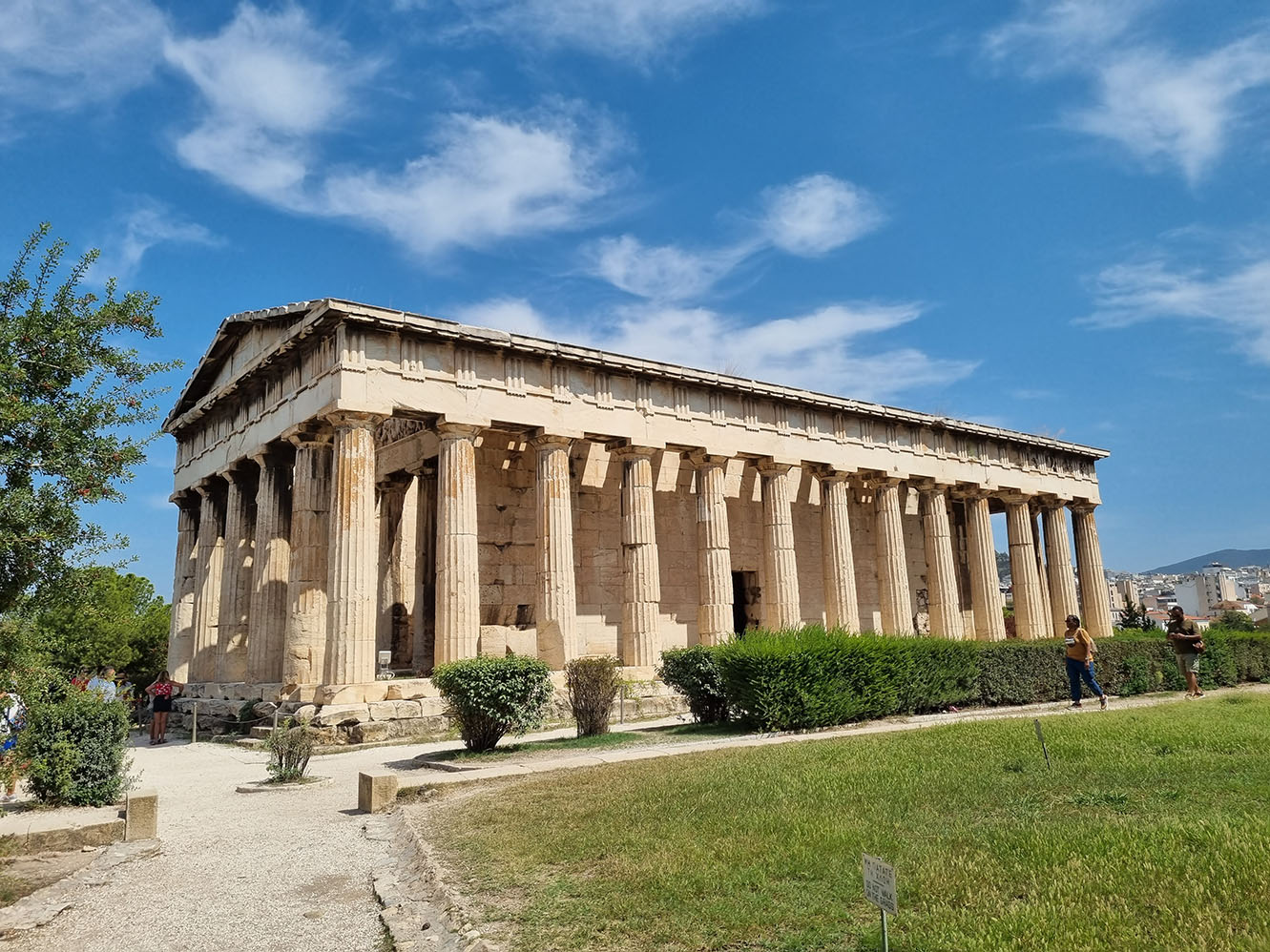 Der Tempel des Hephaistos in Athen, Griechenland, mit dorischen Säulen und intaktem Dach, unter einem klaren blauen Himmel, mit einer vorbeigehenden Person und viel Grün drumherum.