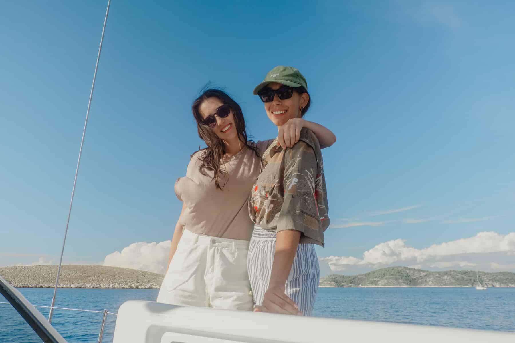 Zwei Frauen in lässiger Sommerkleidung lächeln und posieren zusammen auf einer Segelyacht, mit einem klaren blauen Himmel und ruhigem Meer im Hintergrund.