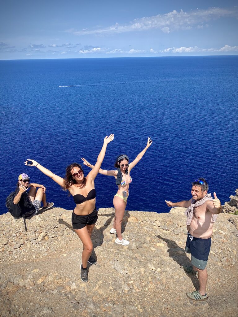 Drei Freunde posieren fröhlich auf einer Felsklippe mit einem weiten blauen Meer und Himmel im Hintergrund. Zwei Frauen und ein Mann feiern mit ausladenden Gesten die atemberaubende Landschaft um sie herum, einschließlich ihrer nahe gelegenen Segelyacht.