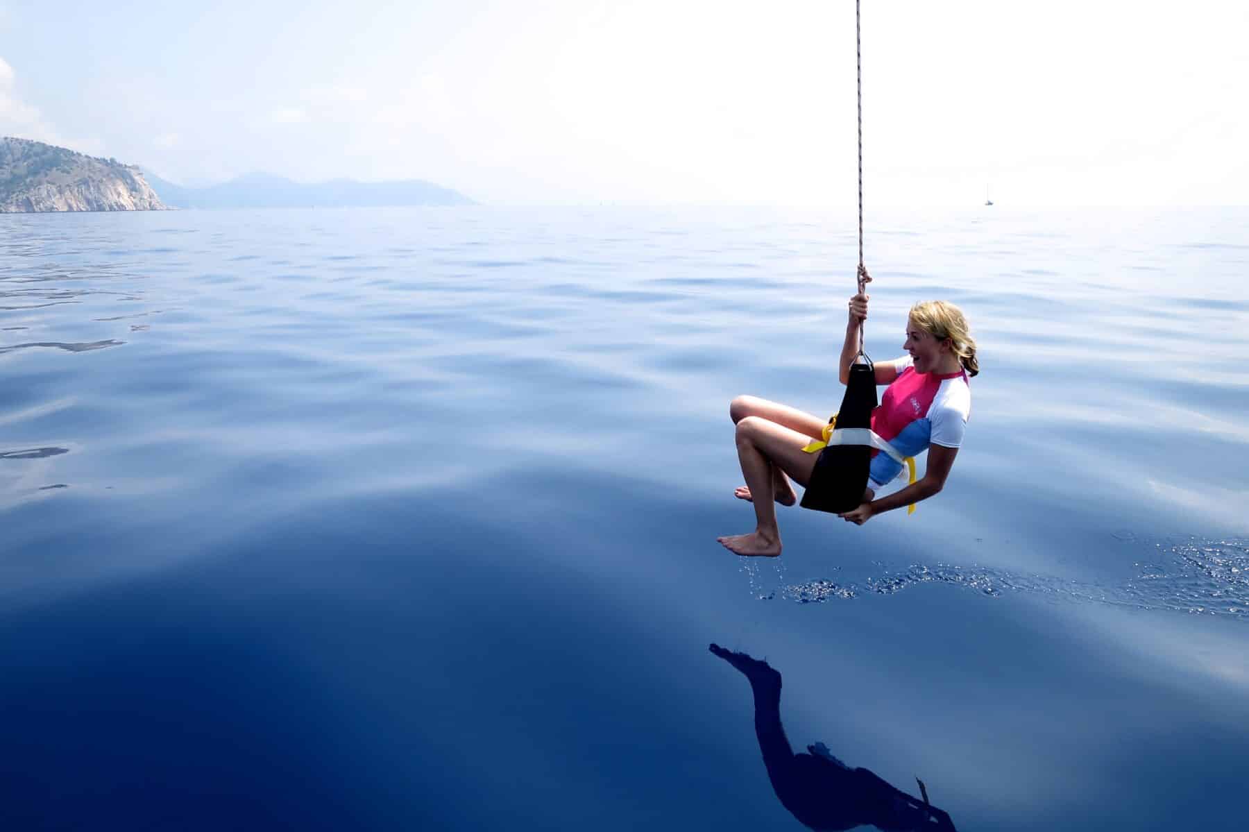 Eine Person in einer Schwimmweste sitzt auf einer Schaukel über dem ruhigen, blauen Ozean, im Hintergrund sind schwach Berge zu erkennen. Der klare Himmel und das ruhige Wasser schaffen während des Segeltörns eine friedliche Szene.