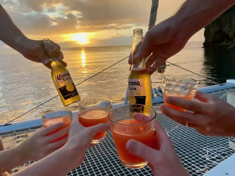 Menschen stoßen bei Sonnenuntergang auf einem Katamaran mit einem orangefarbenen Getränk an, mit Blick auf das Meer und sichtbaren Corona-Bierflaschen.