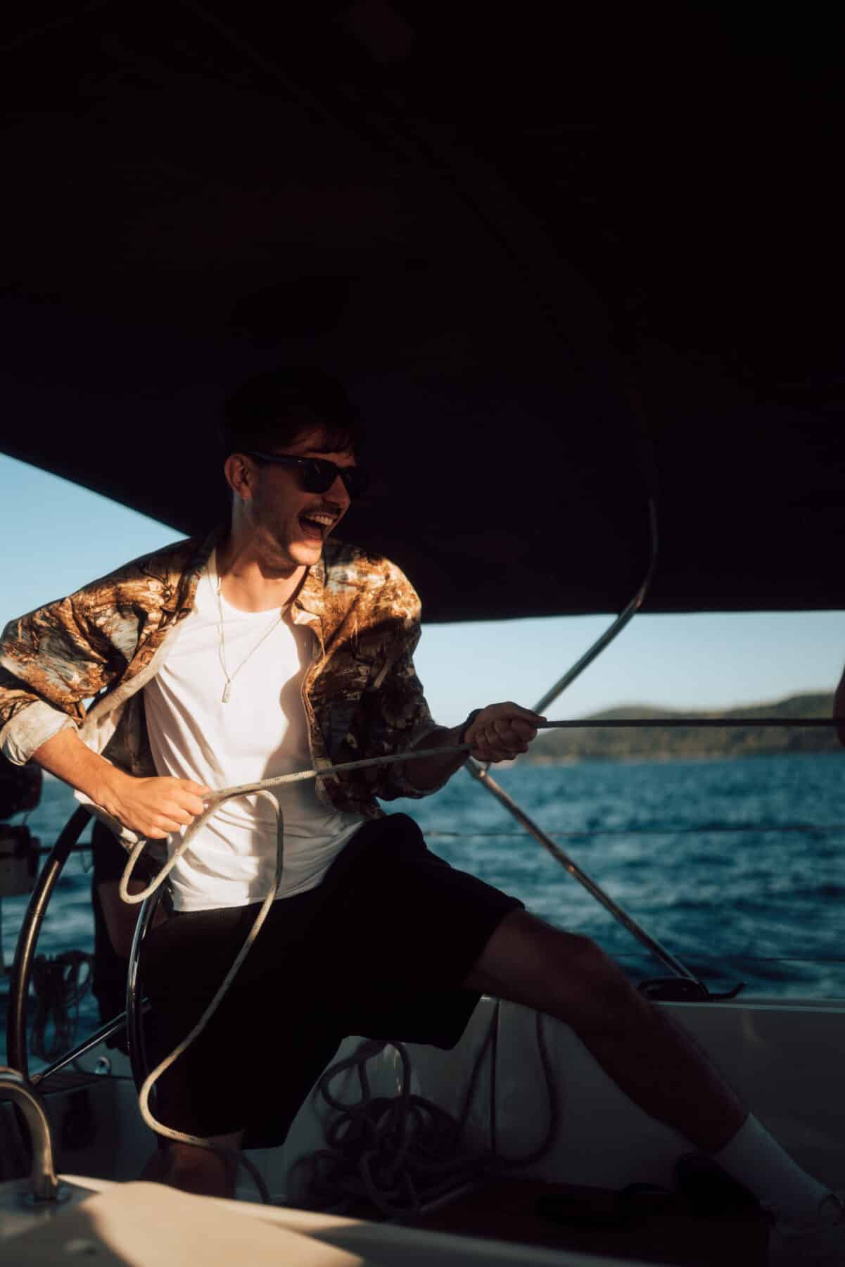 Ein Mann mit Sonnenbrille und geblümtem Hemd lächelt, während er auf seinem Segelurlaub ein sonnendurchflutetes Segelboot vor der Wasserkulisse steuert.