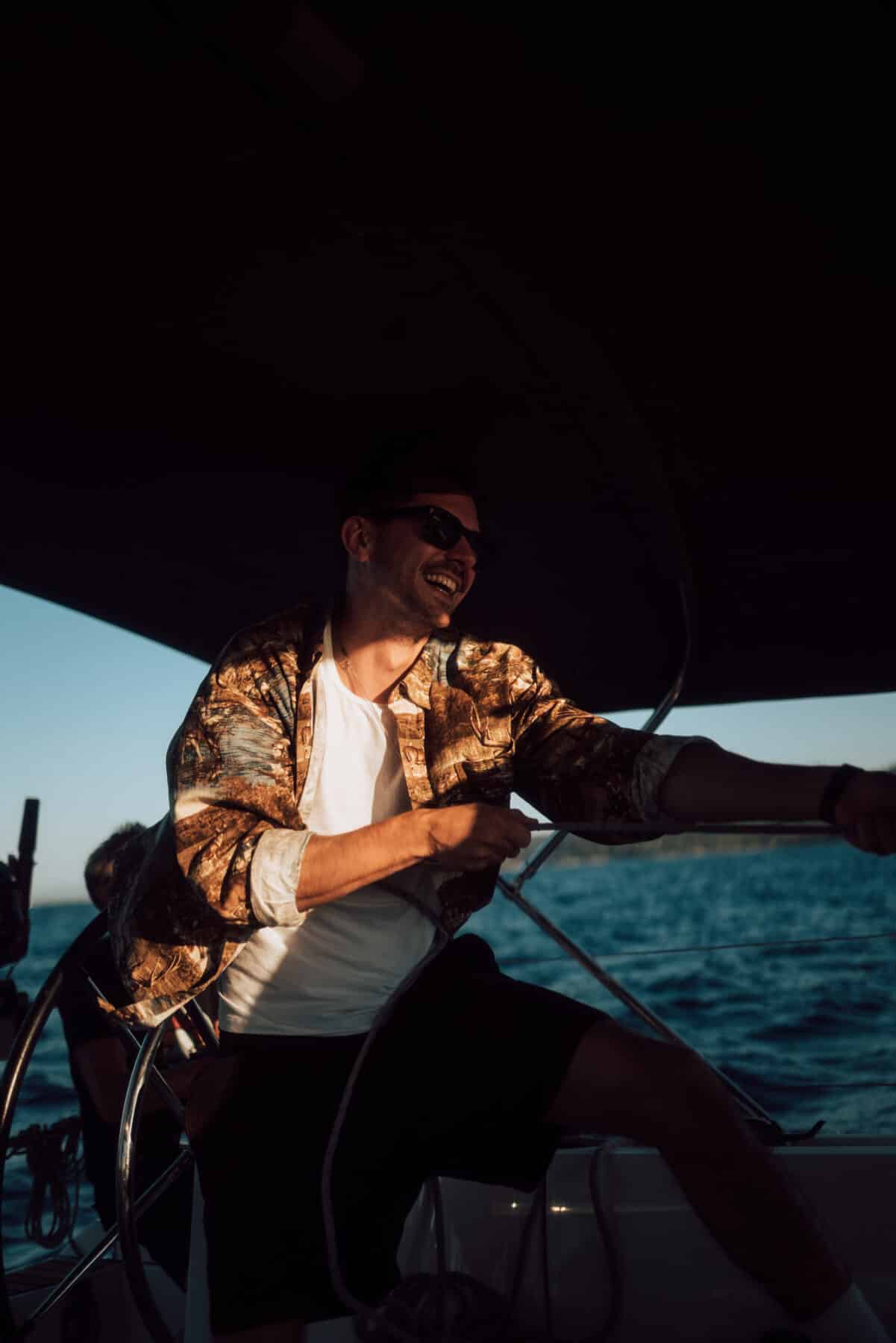 Ein fröhlicher Mann steuert eine Segelyacht. Er trägt eine Sonnenbrille und ein gemustertes Hemd und wird vom Licht der untergehenden Sonne auf dem Wasser beleuchtet.