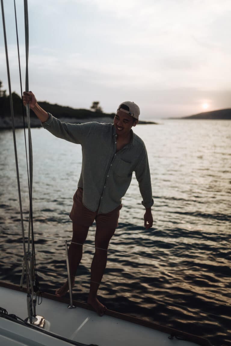 Ein Mann mit Mütze und Freizeitkleidung lächelt, während er sich bei Sonnenuntergang an der Takelage eines Segelboots festhält, mit ruhigem Meer und Hügeln im Hintergrund während seines Segeltörns.