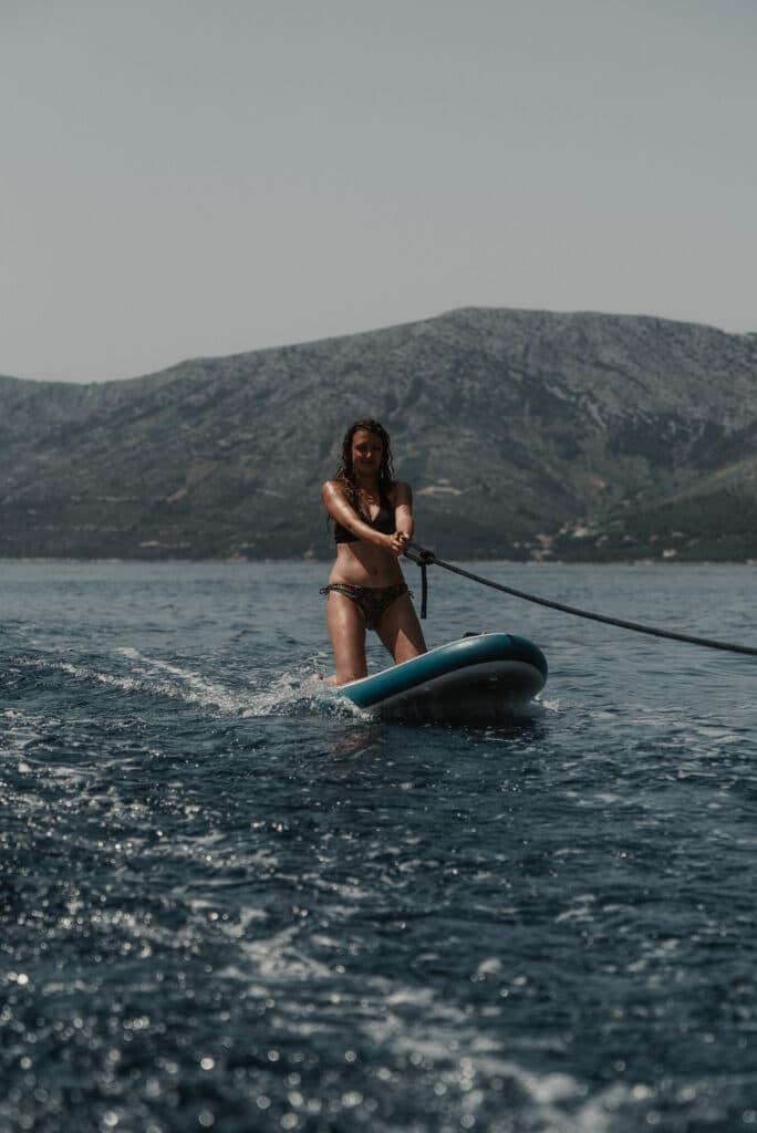 Eine Frau im Bikini macht während ihres Segelurlaubs auf dem Kneeboard eine ruhige See mit Bergen im Hintergrund und nutzt ein Schleppseil, um das Gleichgewicht zu halten.