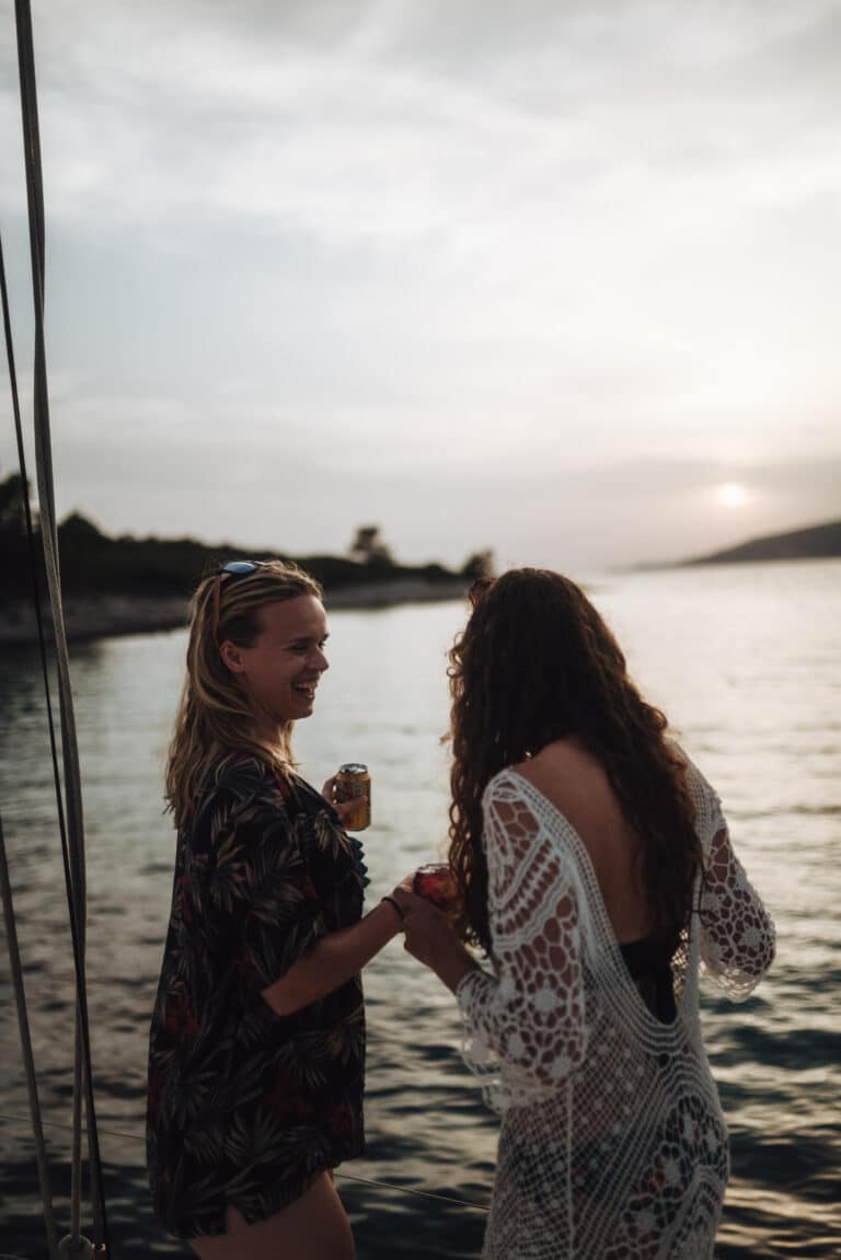 Zwei Frauen lachen und halten Drinks auf einer Segelyacht bei Sonnenuntergang, mit ruhigem Wasser und fernen Ufern im Hintergrund.