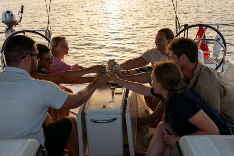Eine Gruppe von Freunden genießt bei Sonnenuntergang auf einem Katamaran Getränke und lacht gemeinsam, während sich das goldene Sonnenlicht im Meer spiegelt.