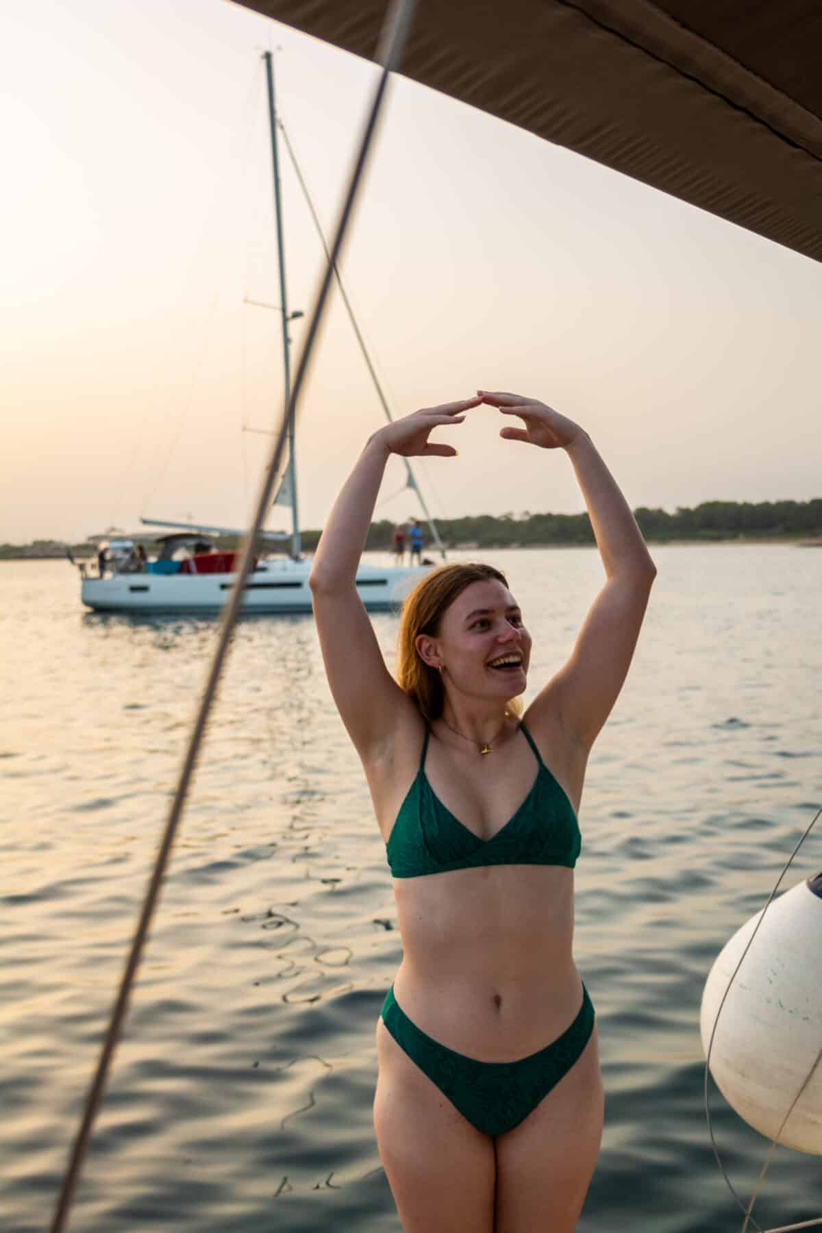 Eine fröhliche Frau in einem grünen Bikini formt mit den Händen über dem Kopf ein Herz und steht bei Sonnenuntergang an einem Seeufer mit Segelbooten und Katamaranen im Hintergrund.