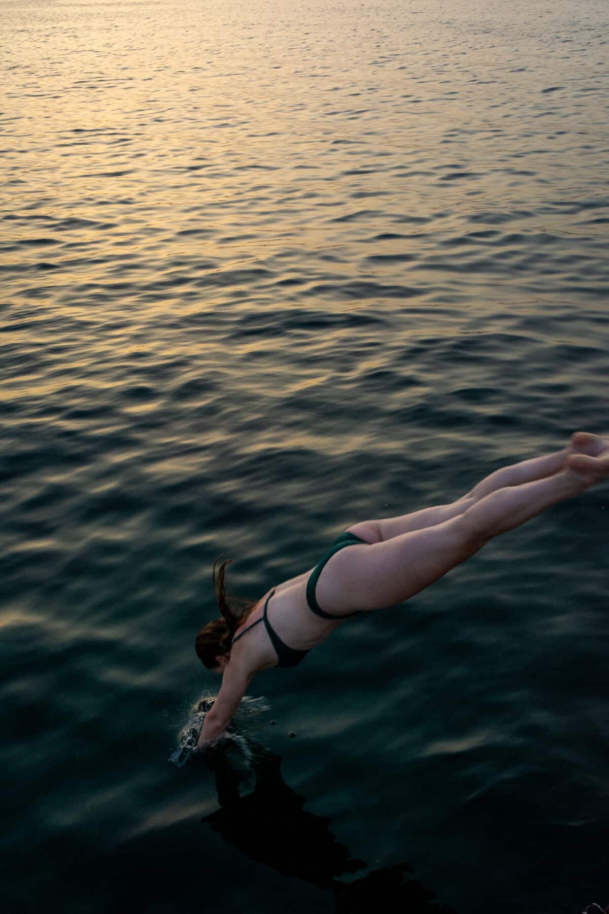 Eine Person taucht bei Sonnenuntergang von einer Segelyacht in ein ruhiges Meer ein, wobei sich ihr Körper in perfekt ausgerichteter, stromlinienförmiger Position knapp über der Wasseroberfläche befindet.