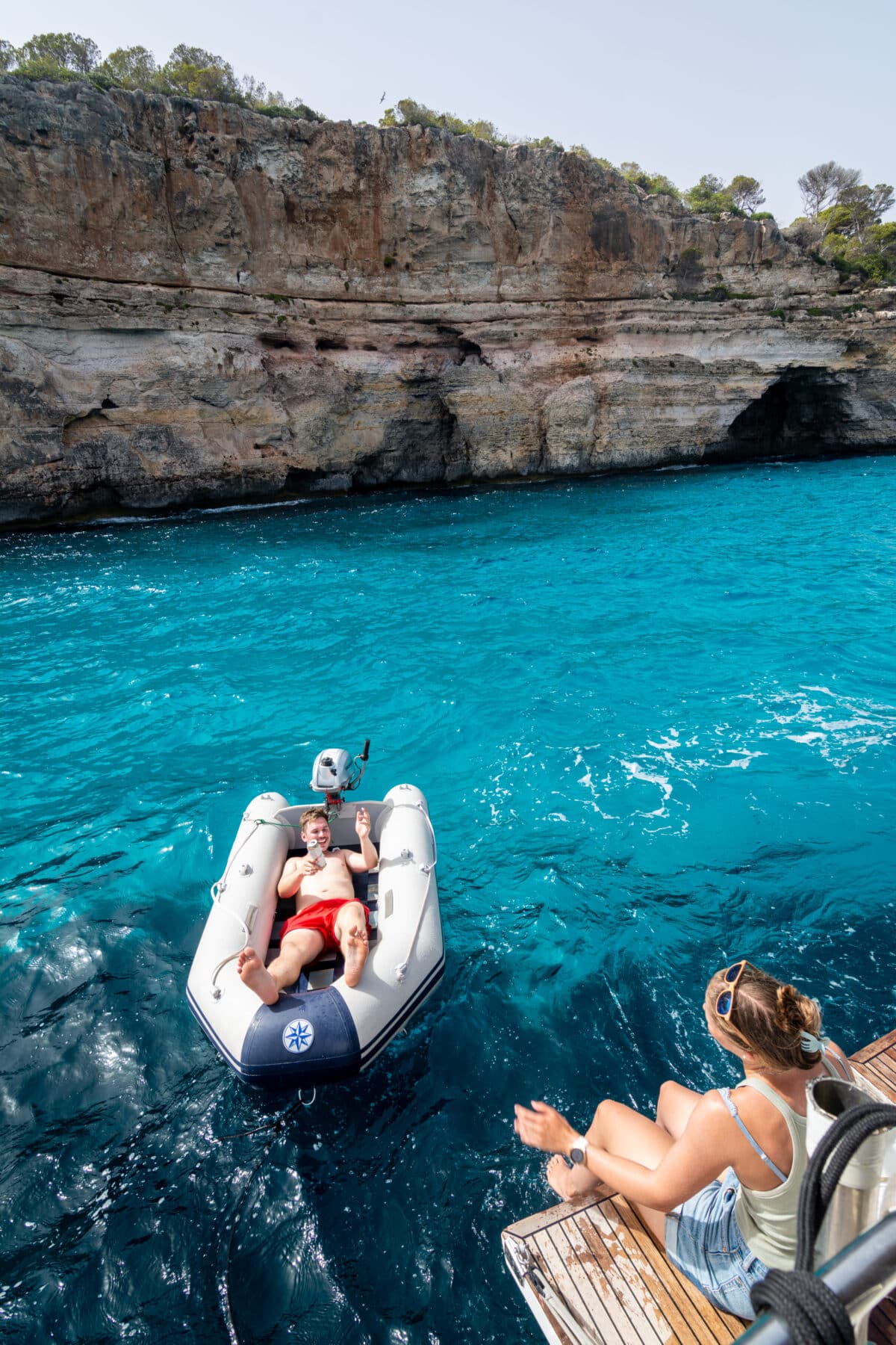 Eine Frau sitzt auf einer Bootsplattform, lässt ihre Beine in Richtung Wasser baumeln und winkt zwei lächelnden Personen in einem kleinen Schlauchboot zu. Sie treiben im hellen Sonnenlicht auf klarem, türkisfarbenem Wasser in der Nähe einer Felsklippe. Auf der Klippe wachsen Bäume und Sträucher – eine idyllische Kulisse für ihr Segeltörn-Abenteuer.