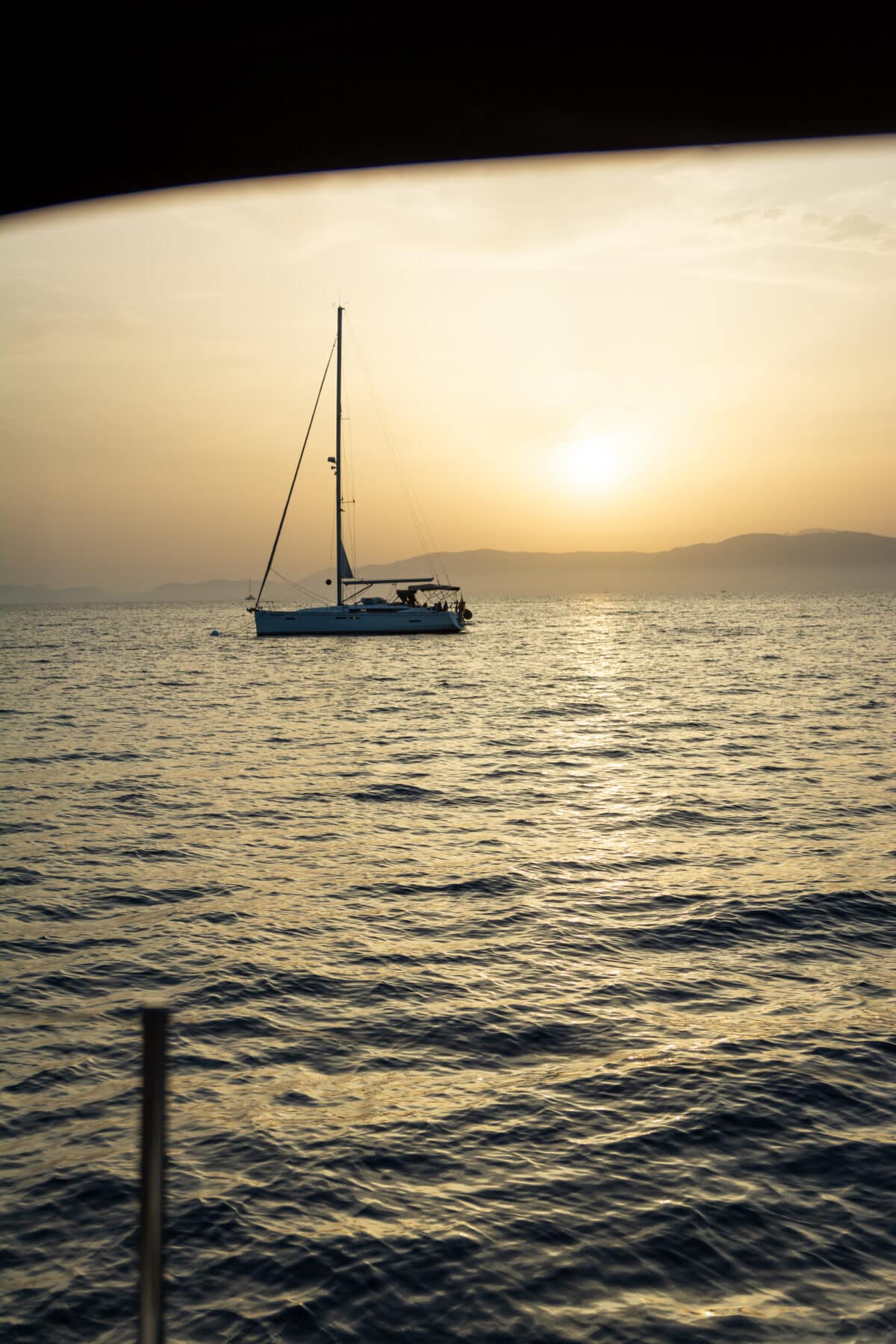 Ein einzelner Katamaran schwimmt bei Sonnenuntergang auf ruhigem Wasser. Der Himmel ist golden und die Sonne steht tief und wirft ein warmes Spiegelbild auf das Wasser. Im Hintergrund sind die Silhouetten der fernen Berge zu sehen, die die perfekte Kulisse für einen ruhigen Segelurlaub schaffen.