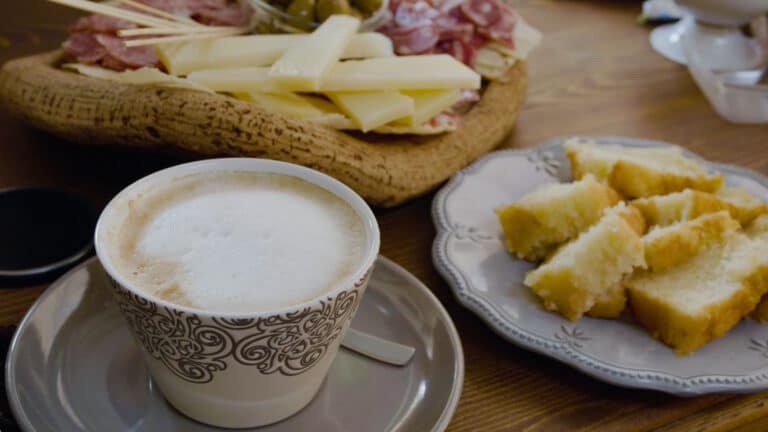 Eine ruhige Kaffeetafel mit einer Tasse Cappuccino mit einer dicken Schaumschicht, einem Teller mit geschnittenem Brot und einem Korb voller Käsesorten und Aufschnitt auf einem Katamaran.