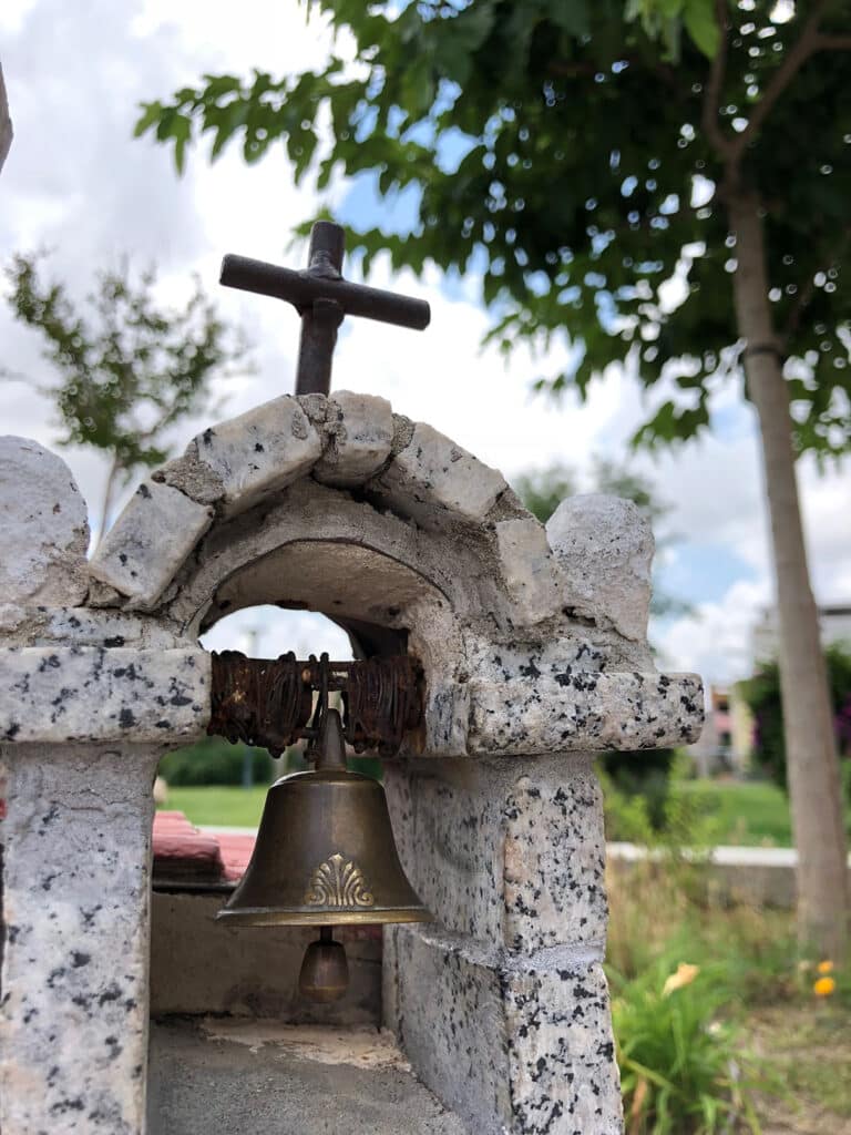 Ein kleiner, rustikaler Steinschrein mit einem Bogen, auf dem sich ein Kreuz befindet und in dem eine Glocke hängt, vor einem verschwommenen Hintergrund aus grünen Bäumen und einem Katamaran.