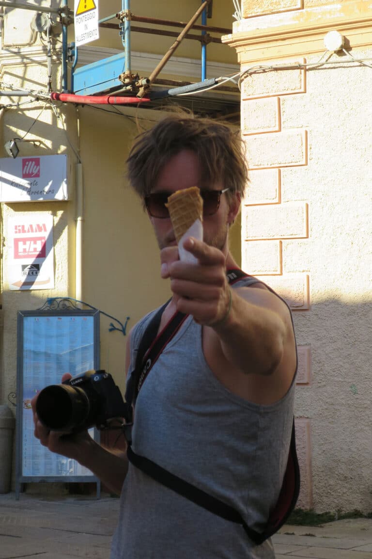 Ein Mann mit zerzaustem Haar hält eine Eistüte in die Kamera, während er eine DSLR-Kamera trägt. Er steht auf einem sonnenbeschienenen Katamaran mit Gebäuden und Schildern im Hintergrund.