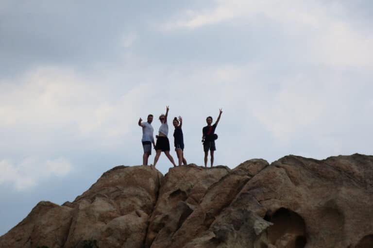Drei Menschen stehen auf einer großen Felsformation, winken und strecken ihre Arme vor einem bewölkten Himmel.