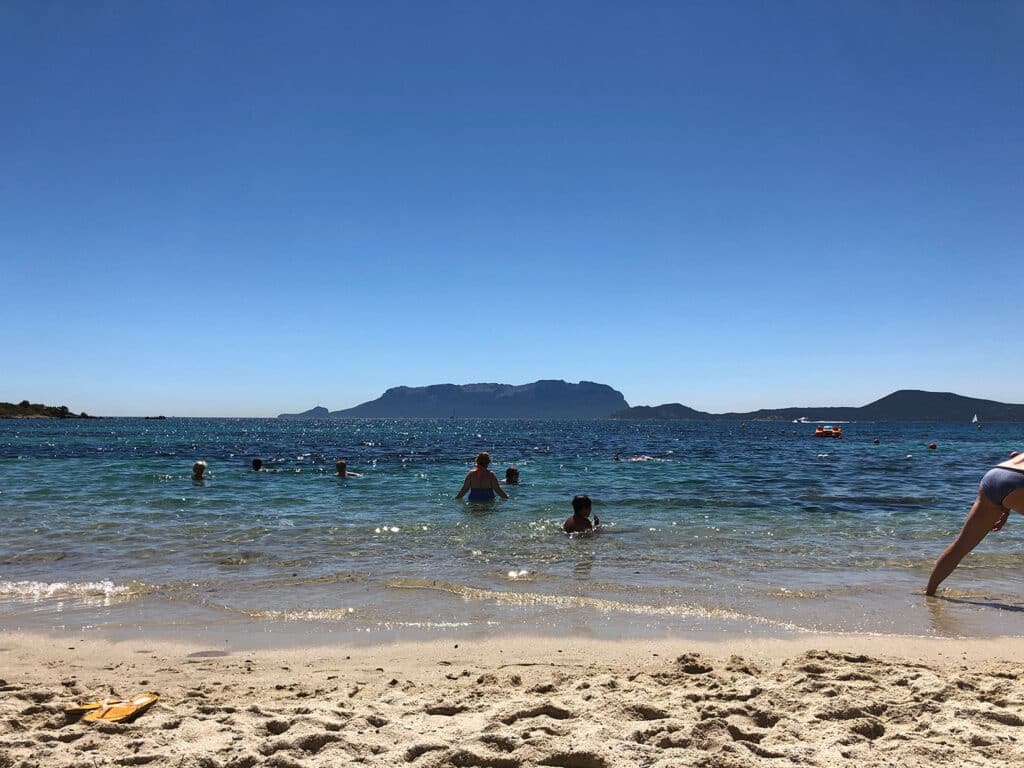 Eine sonnige Strandszene mit Menschen, die im Meer schwimmen und im Sand entspannen. In der Ferne segelt ein Katamaran in der Nähe eines Berges, der unter einem klaren blauen Himmel aus dem Meer ragt.