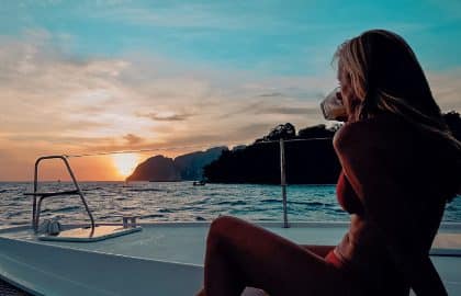 Eine Frau in einem roten Kleid sitzt auf einem Katamaran und blickt auf den Sonnenuntergang über dem Meer, im Hintergrund ist die Silhouette einer Insel zu sehen.