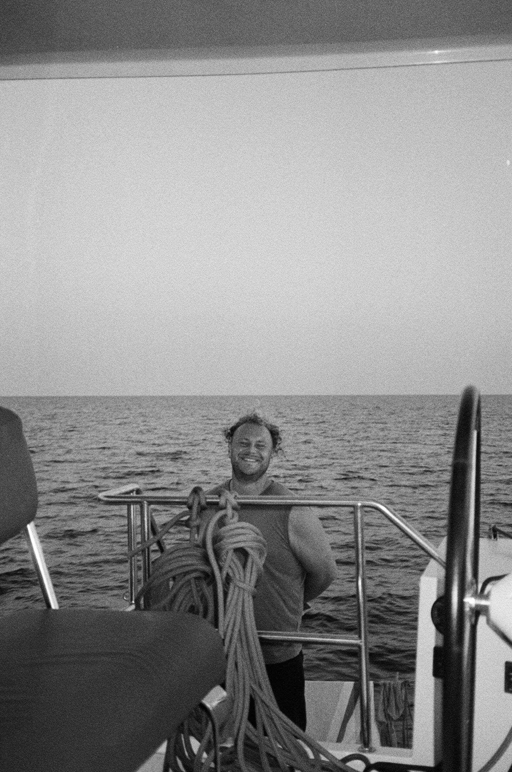 Eine Person steht lächelnd und in einem Tanktop auf einem Boot. Sie steht hinter einem Geländer mit aufgerollten Seilen im Vordergrund. Hinter ihnen erstreckt sich der Ozean unter einem klaren Himmel. Das Schwarzweißbild fängt die Essenz ihrer Segelreise auf einem schnittigen Katamaran ein.