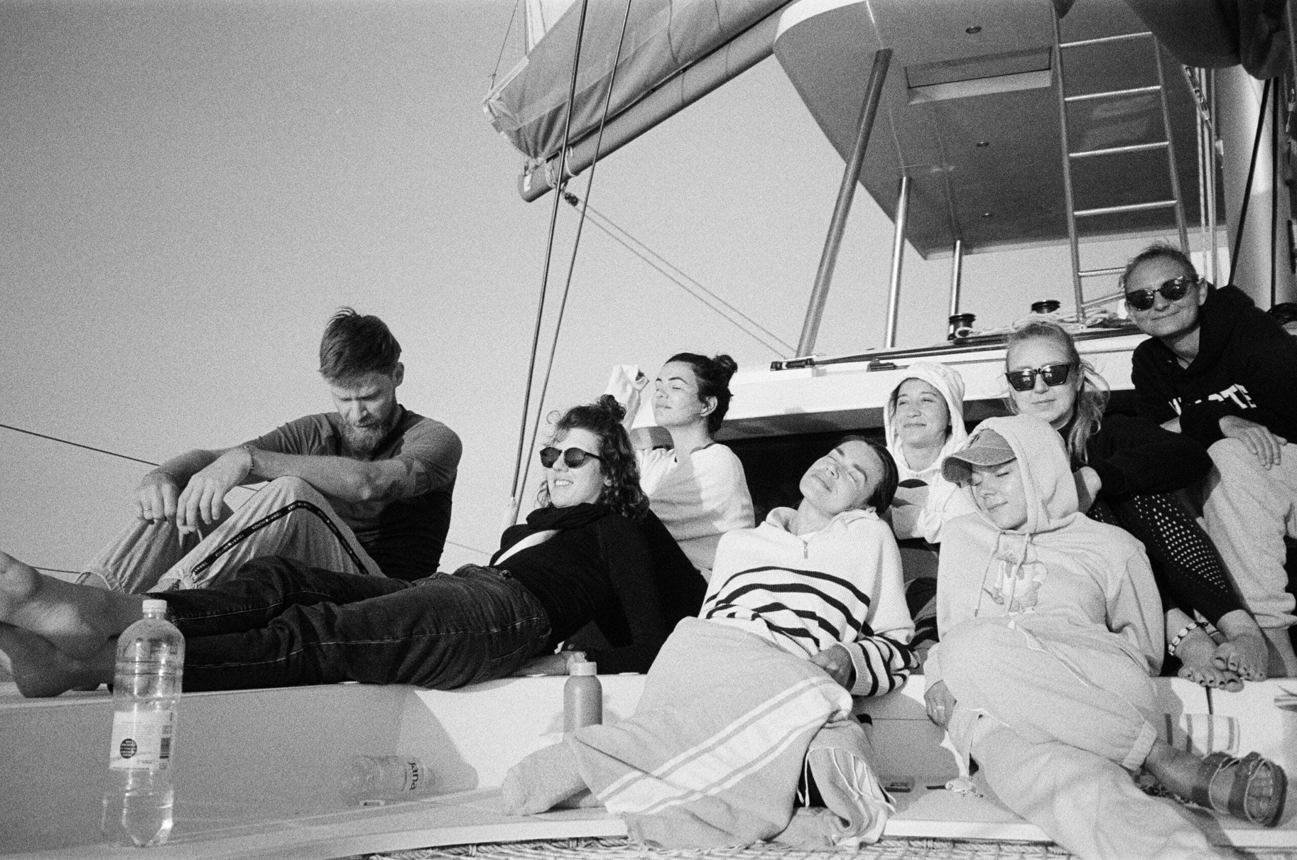 Eine Gruppe von sieben Personen entspannt sich und genießt das Leben auf einer Segelyacht. Sie tragen legere Kleidung und Sonnenbrillen und aalen sich unter einem klaren Himmel.
