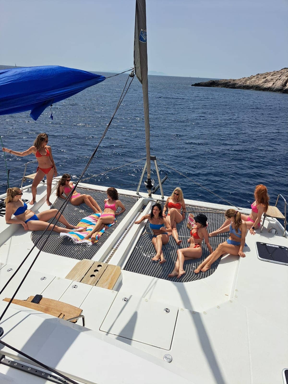 Eine Gruppe von Menschen genießt einen sonnigen Tag auf dem Deck eines Katamarans, umgeben von klarem, blauem Wasser, wobei einige sitzen und andere stehen.
