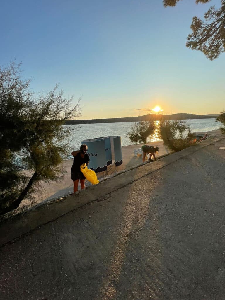 Sonnenuntergang über einem ruhigen See mit Menschen, die am Ufer entlanggehen, einige tragen Gegenstände. Bäume beschatten den Weg teilweise und eine helle Sonne schwebt über dem Horizont. Ein Katamaran segelt sanft am Wasser entlang.