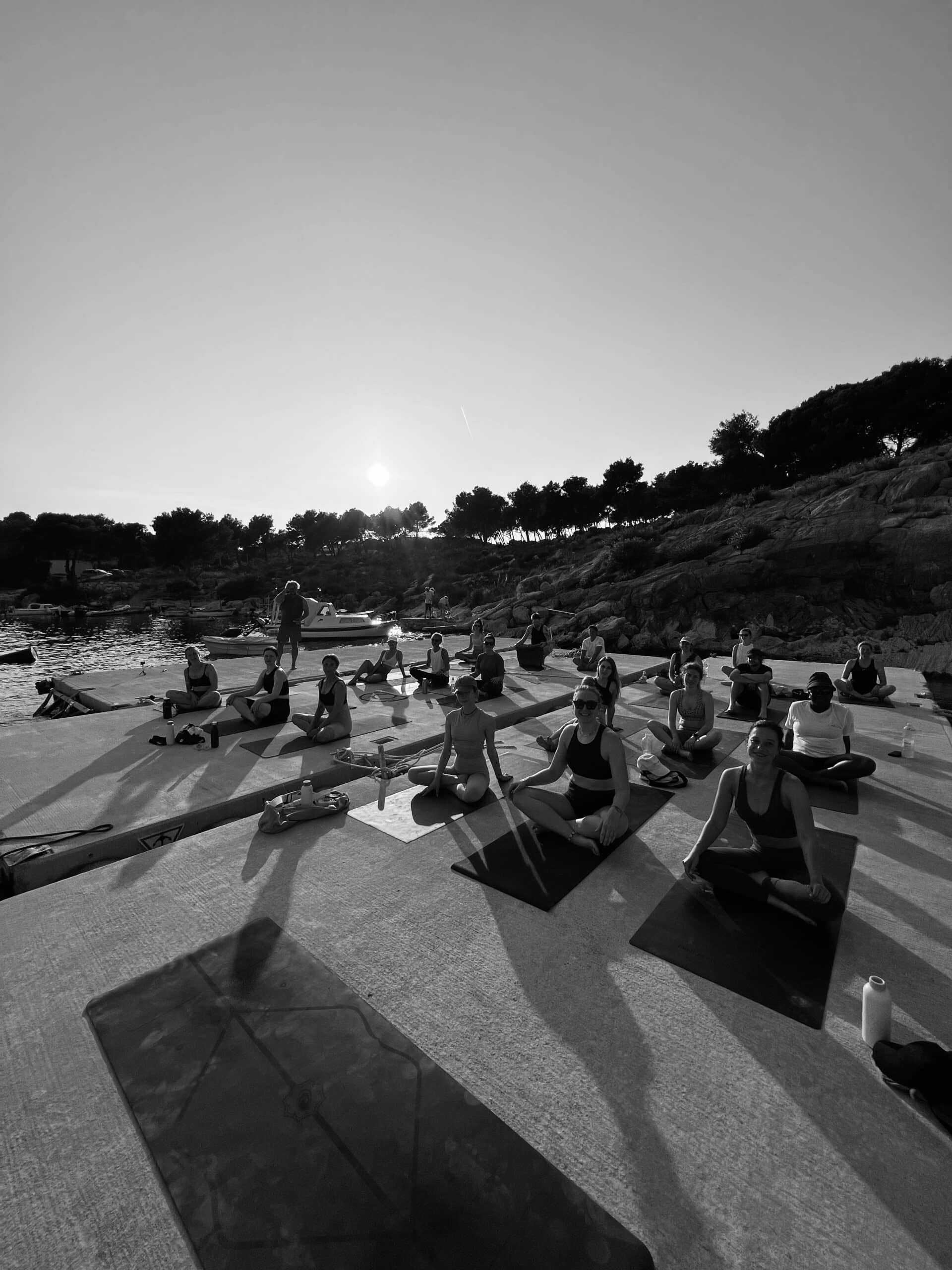 Ein Schwarzweißfoto einer Gruppe, die während ihres Segelurlaubs in einer natürlichen, felsigen Umgebung unter klarem Himmel auf Matten Yoga praktiziert, im Hintergrund die Sonne untergeht.