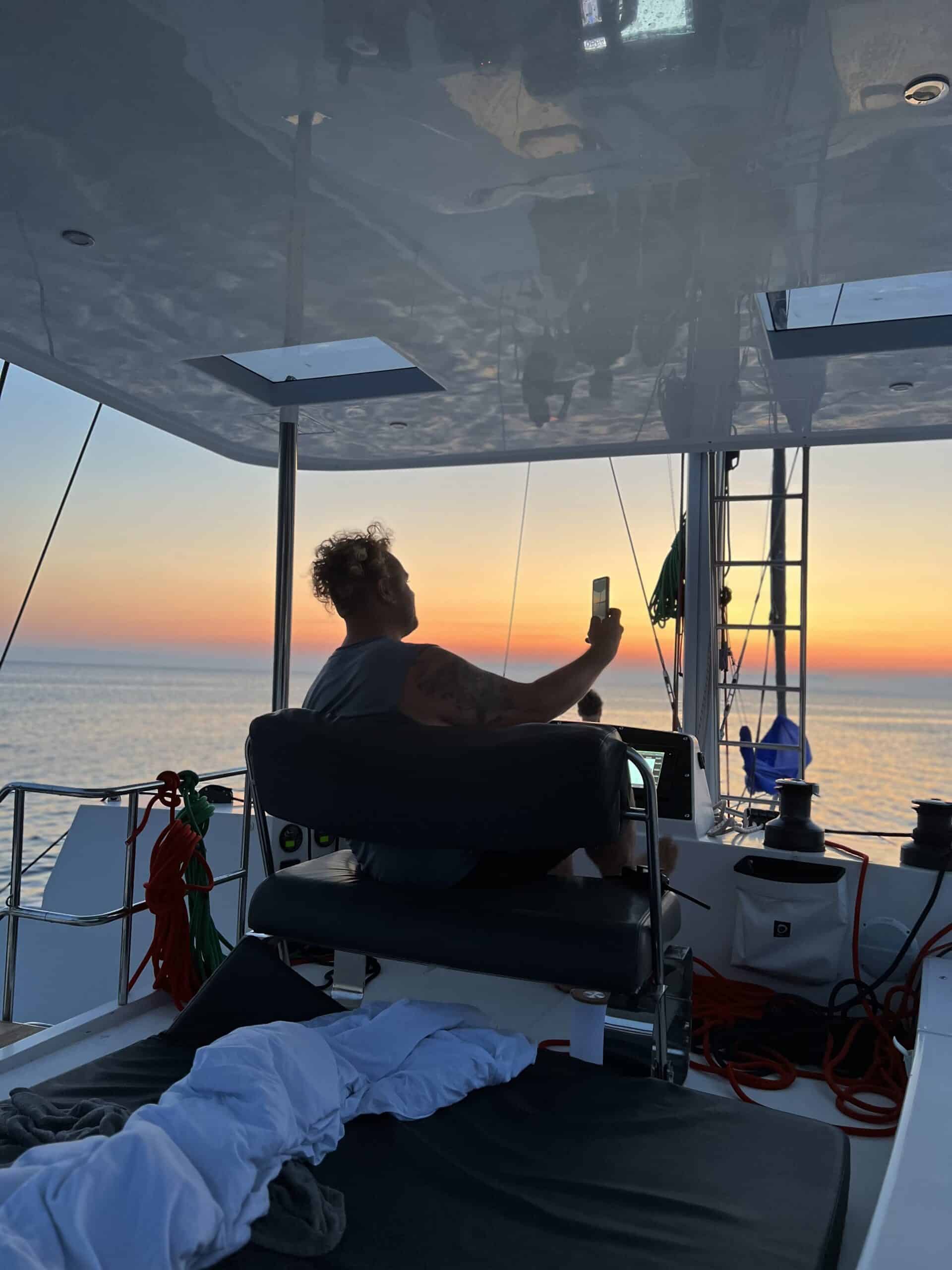 Eine Person sitzt bei Sonnenuntergang auf einer Segelyacht und hält ein Mobiltelefon hoch, um die Szene festzuhalten. Im Hintergrund erstreckt sich der Ozean unter einem Himmel mit Farbverlauf.