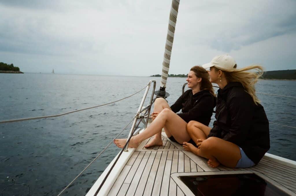 Zwei Frauen sitzen auf dem Deck eines Katamarans und blicken auf das Wasser, mit einem bewölkten Himmel über ihnen und einem weiteren Segelboot in der Ferne.