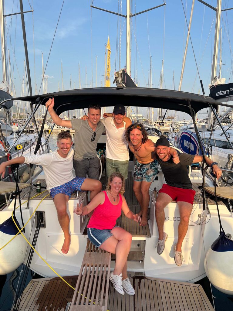 Sechs Freunde posieren glücklich auf dem Heck eines Katamarans in einem sonnigen Yachthafen, vier Männer und eine Frau, alle in legerer Sommerkleidung, und lächeln in die Kamera.