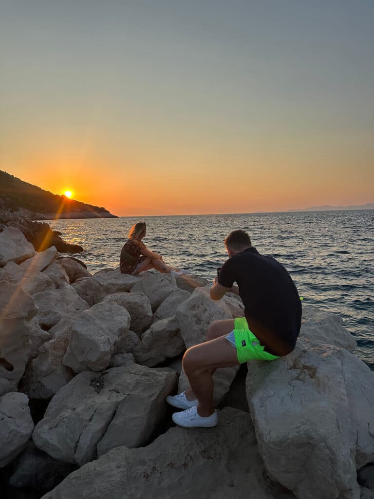 Zwei Personen sitzen auf großen Felsen am Meer und beobachten während ihres Segeltörns einen Sonnenuntergang. Eine Person, die abgewandt ist, hält die Szene mit einer Kamera fest. Die Sonne sinkt nahe dem Horizont unter und verbreitet einen warmen Schein.