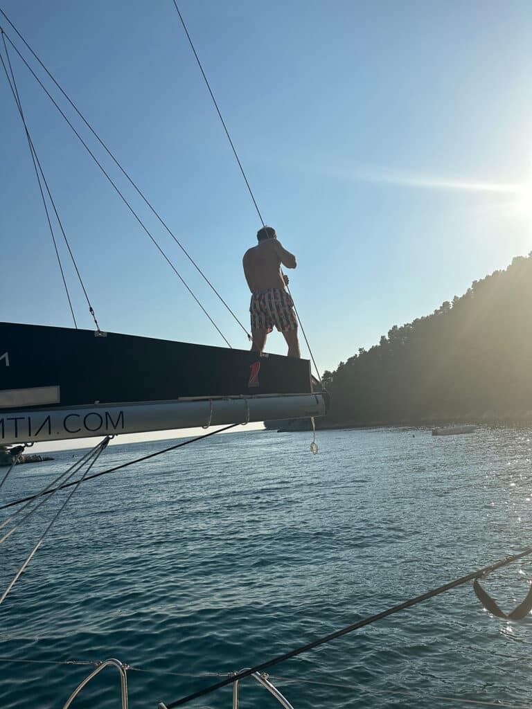 Eine Person mit Hut und gemusterten Shorts steht auf dem Bug eines Segelboots und blickt über das Meer auf eine bewaldete Küste, die während des Segeltörns vom Sonnenlicht beleuchtet wird.