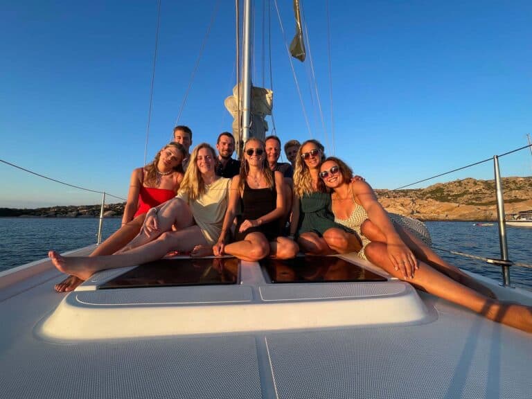 Eine Gruppe von acht Freunden posiert während ihrer Segelreise bei Sonnenuntergang auf dem Vorderdeck eines Segelboots und lächelt freudig vor der malerischen Kulisse des ruhigen Meeres und der felsigen Küste.