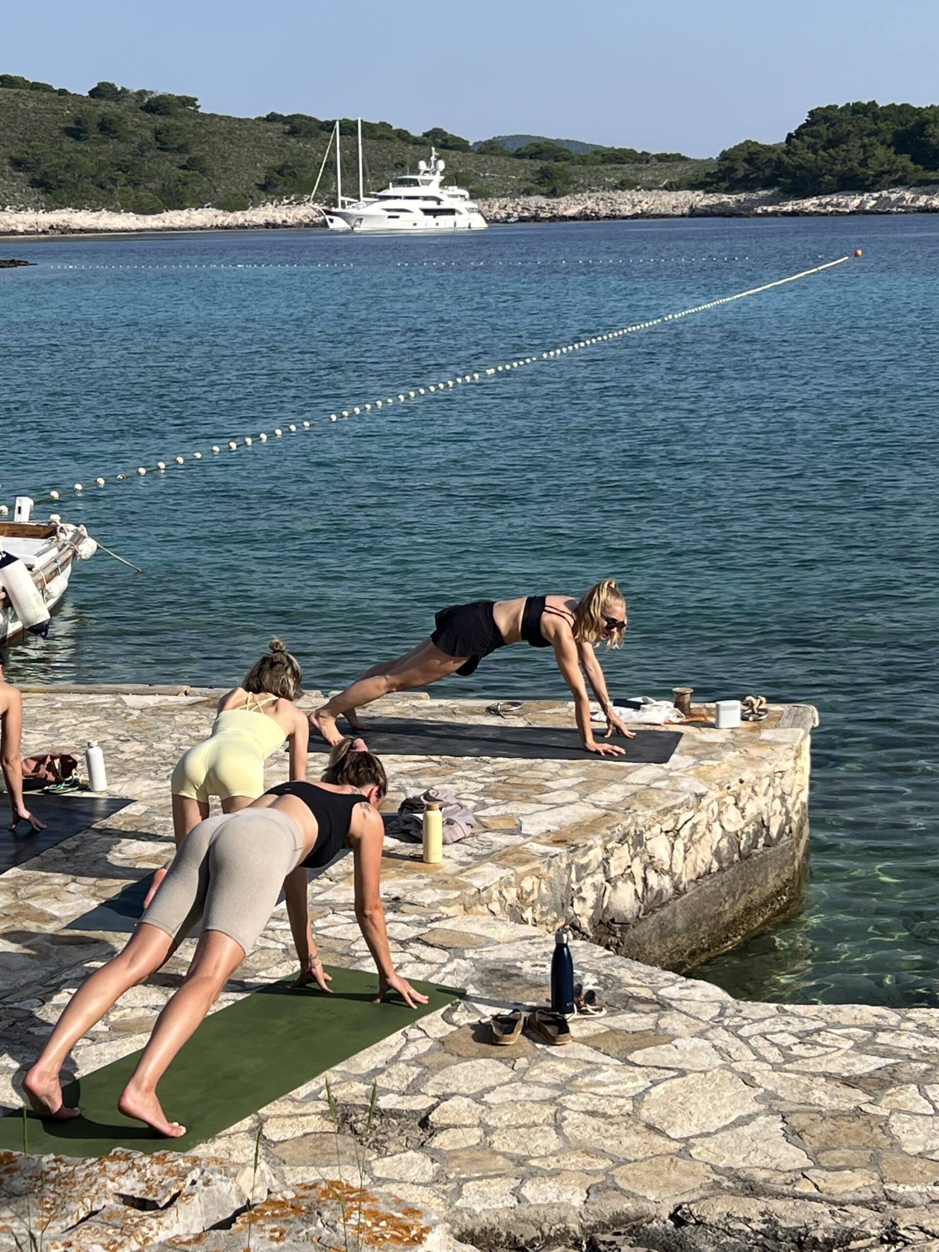Drei Personen praktizieren Yoga auf einem Steinsteg am Meer, im Hintergrund ist eine Segelyacht vor Anker. Sie dehnen sich in der herabschauenden Hundestellung.