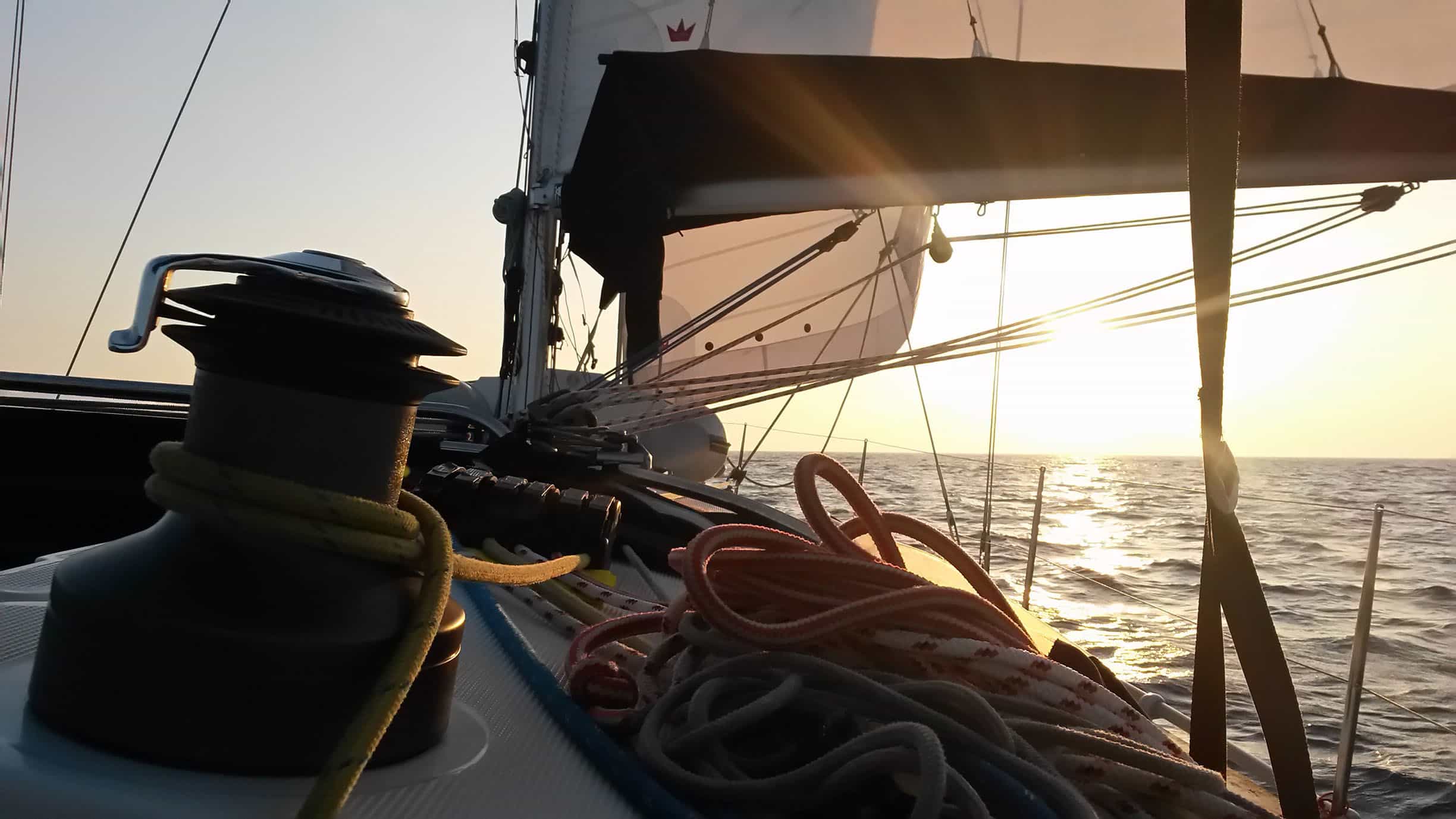 Eine Nahaufnahme des Decks eines Segelboots bei Sonnenuntergang. Zu sehen sind eine Winde mit aufgerollten Seilen und das Großsegel, das während eines Segeltörns vor einem warmen, glühenden Himmel entrollt ist.