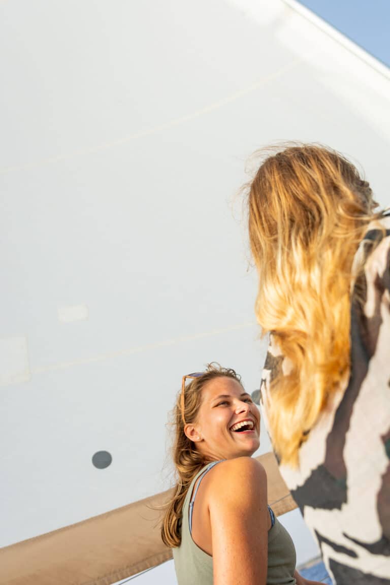 Zwei Frauen unterhalten sich und lachen fröhlich unter einem Segel an einem sonnigen Tag während ihres Segeltörns, eine mit dem Rücken zu ihr, die andere mit einem strahlenden Lächeln in die Kamera blickend.