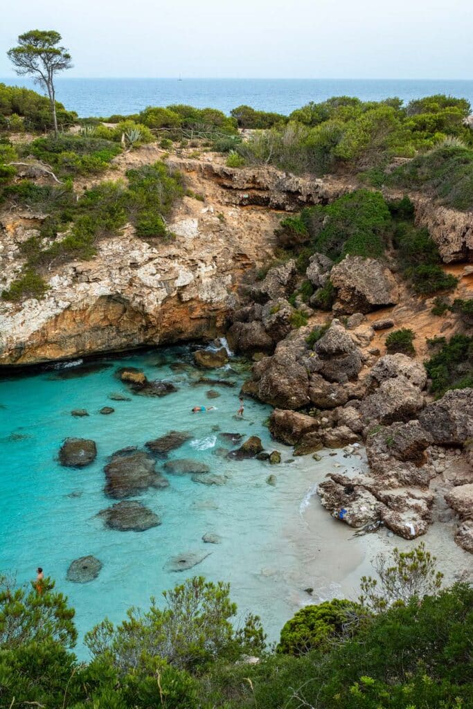 Eine versteckte Bucht mit türkisfarbenem Wasser, umgeben von schroffen Klippen und viel Grün, wo die Leute schwimmen und den abgeschiedenen Strandbereich von Segeltörn genießen.