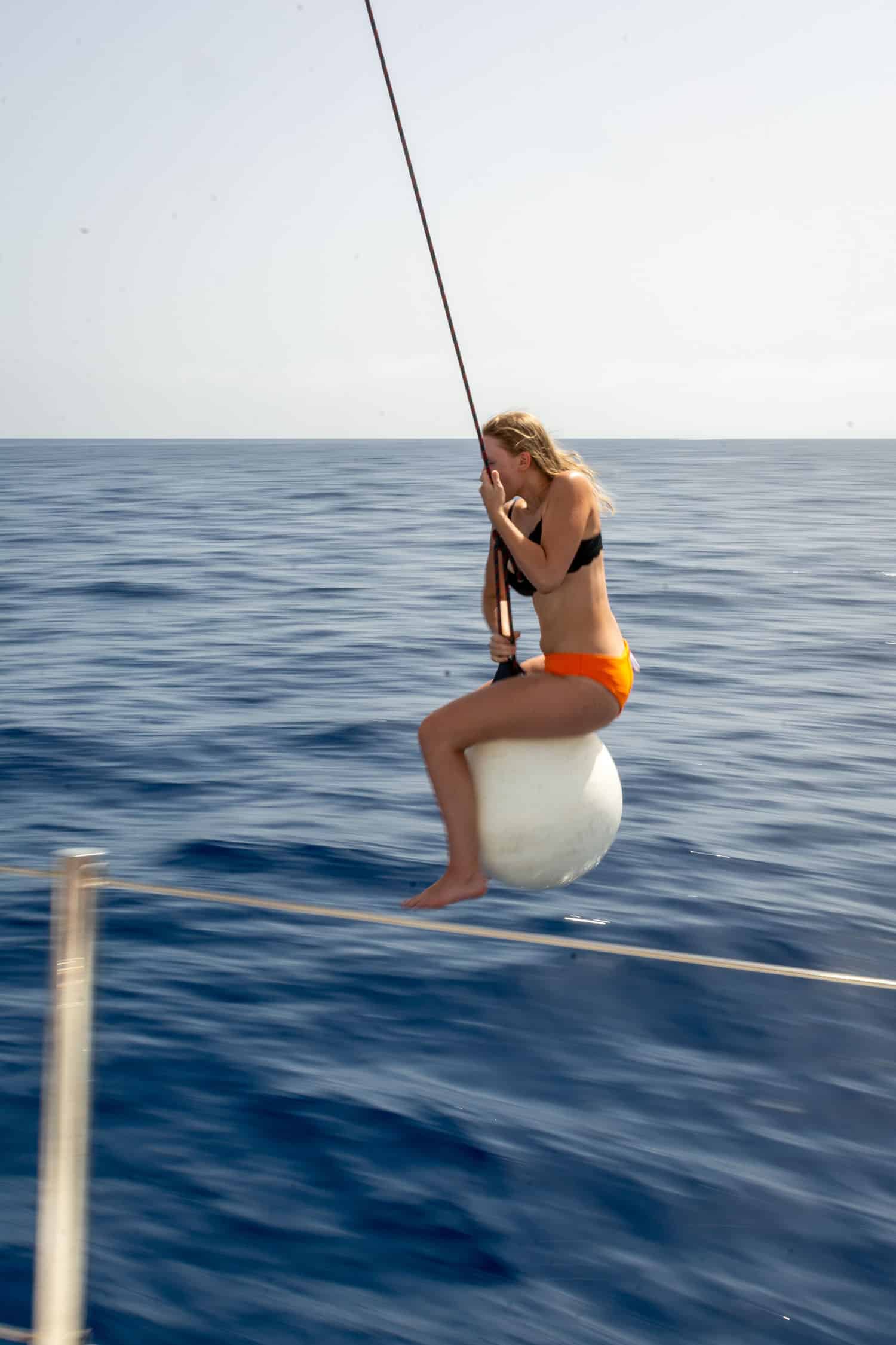Eine junge Frau im orangefarbenen Bikini sitzt auf einer an einer Stange befestigten weißen Boje über dem tiefblauen Meer und konzentriert sich während ihres Segelurlaubs auf das Angeln.