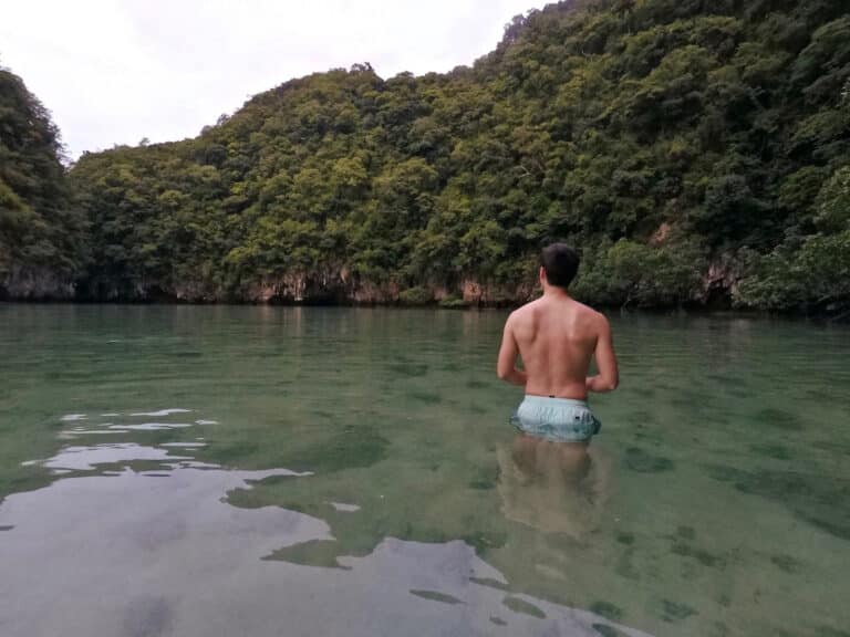 Ein Mann steht hüfttief im ruhigen, türkisfarbenen Wasser und blickt auf die üppigen, grünen Hügel, die das Wasser umgeben. Er ist oben ohne, trägt blaue Shorts und blickt vom Betrachter ab, nachdem er von einer Segelyacht ausgestiegen ist.