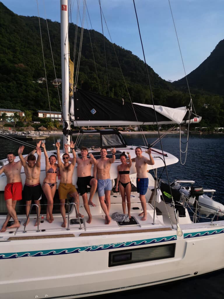 Eine Gruppe glücklicher Freunde in Badeanzügen feiert auf einer Segelyacht mit erhobenen Armen. Das Boot liegt unter einem klaren Himmel in der Nähe einer üppigen, bergigen Küste vor Anker.