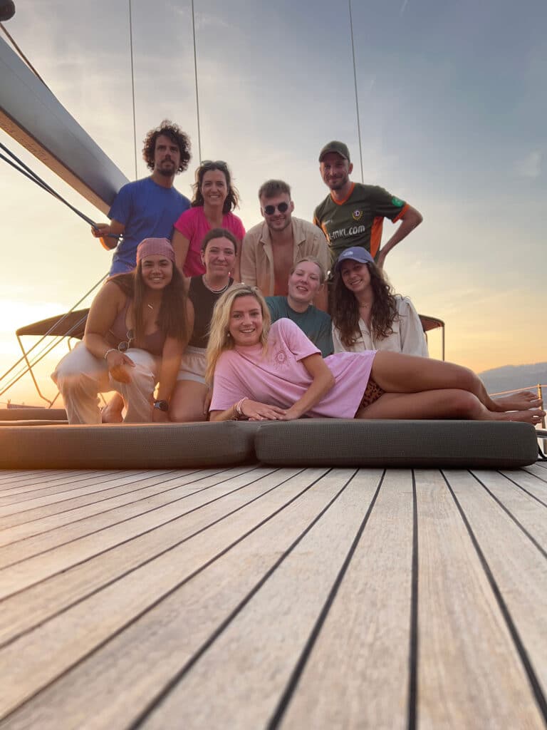 Eine Gruppe von neun jungen Erwachsenen lächelt bei Sonnenuntergang auf dem Deck einer Segelyacht, im Hintergrund ein klarer Himmel. Sie wirken fröhlich und sind leger gekleidet, genießen einen Sommerausflug.