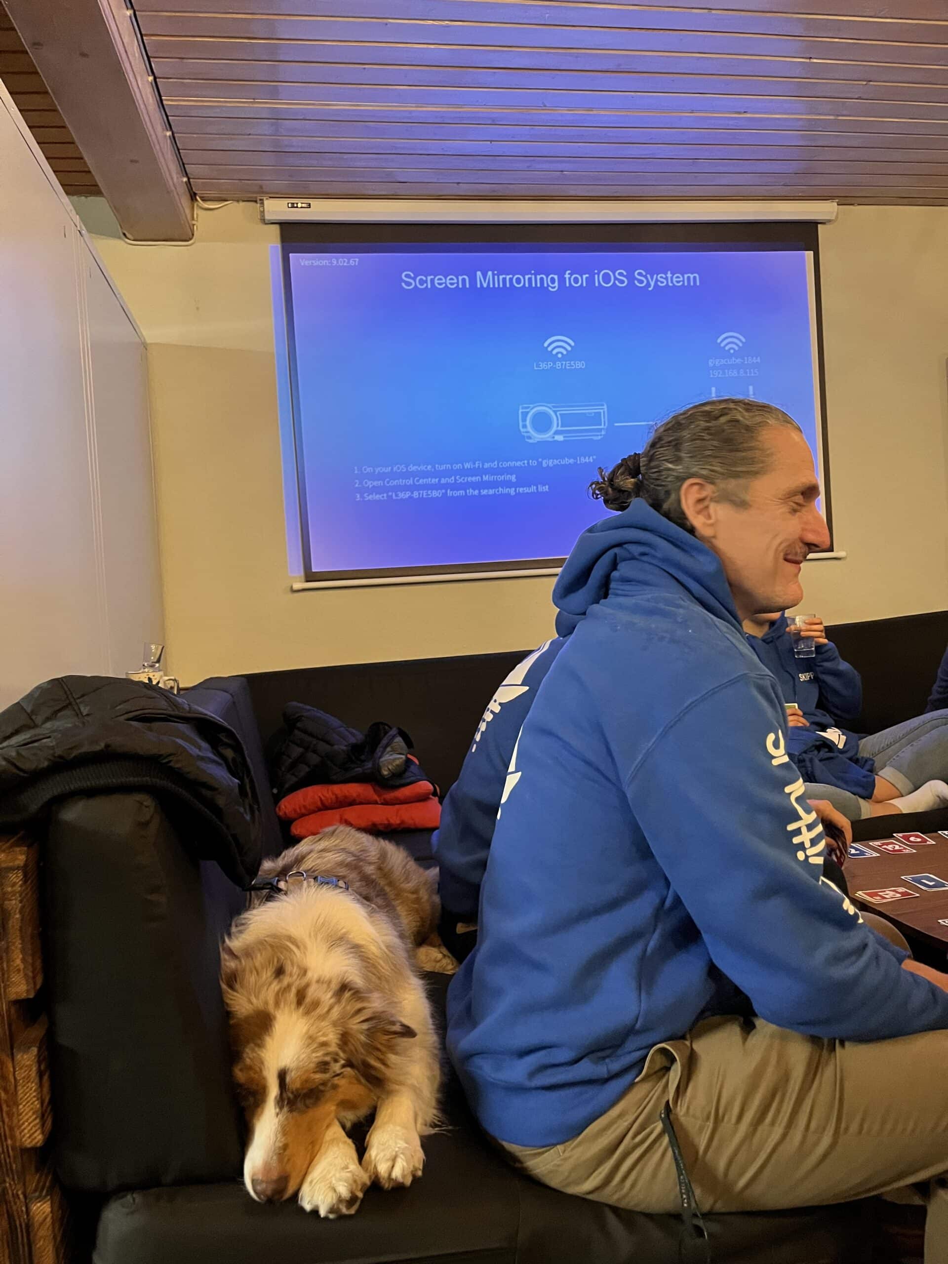 Ein Mann mit einem blauen Kapuzenpullover und ein Shetland Sheepdog sitzen auf einem Sofa in einem Raum mit einem Präsentationsbildschirm, auf dem im Hintergrund „Screen Mirroring für iOS-System“ angezeigt wird.