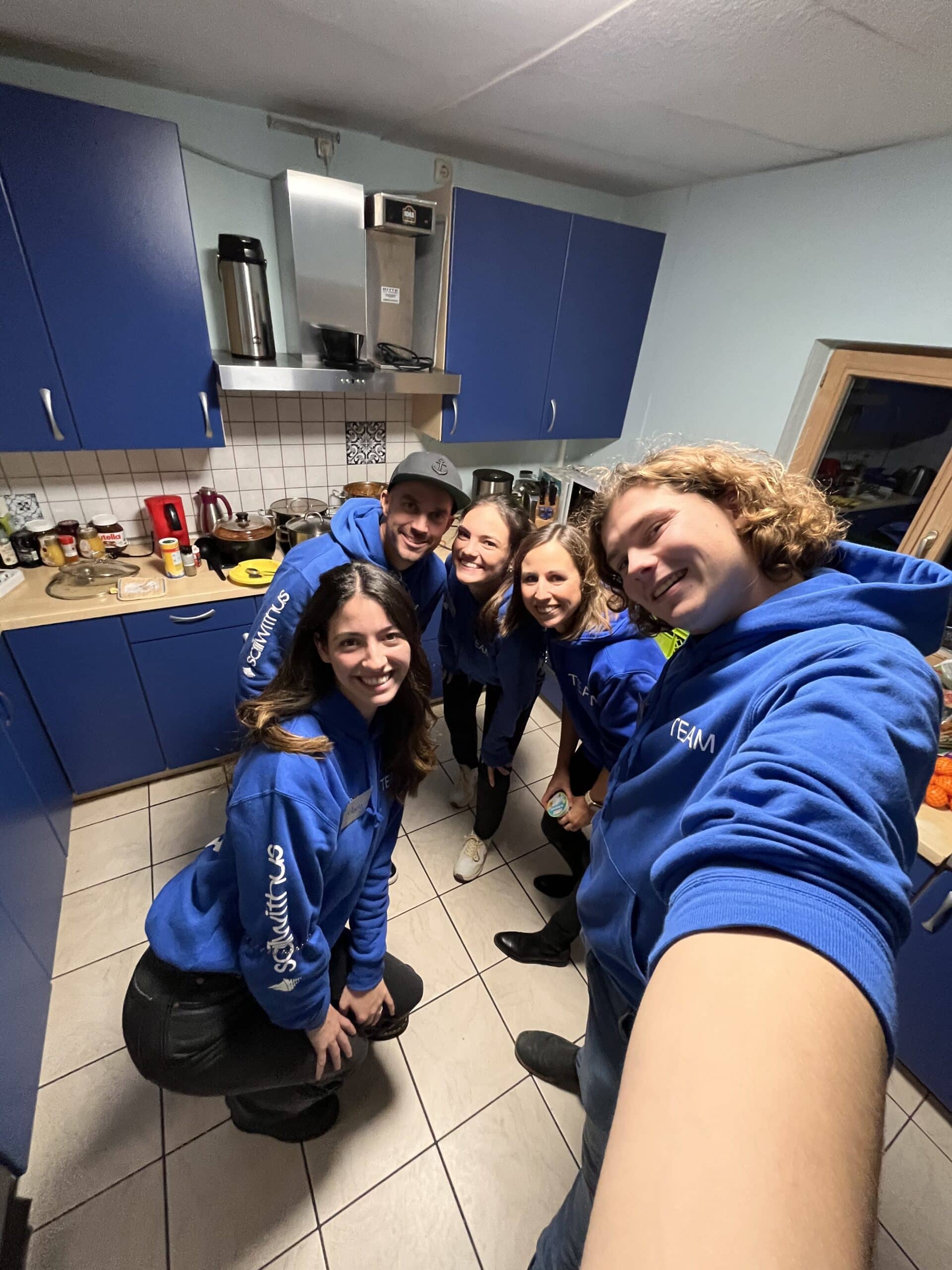 Fünf junge Erwachsene in blauen Kapuzenpullovern lächeln während ihres Segeltörns für ein Gruppen-Selfie in einer Küche mit blauen Schränken und verschiedenen Geräten im Hintergrund.